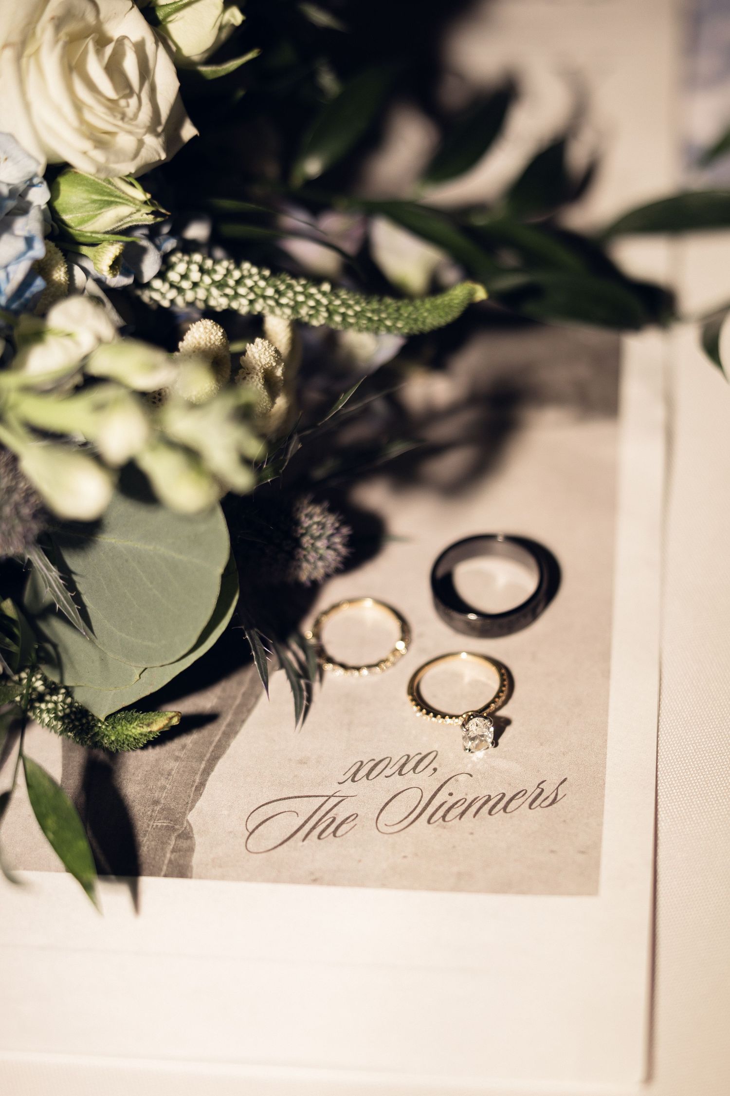 Wedding rings rest on a card signed xoxo, The Siemers, beside white roses, greenery, and delicate blue wildflowers.