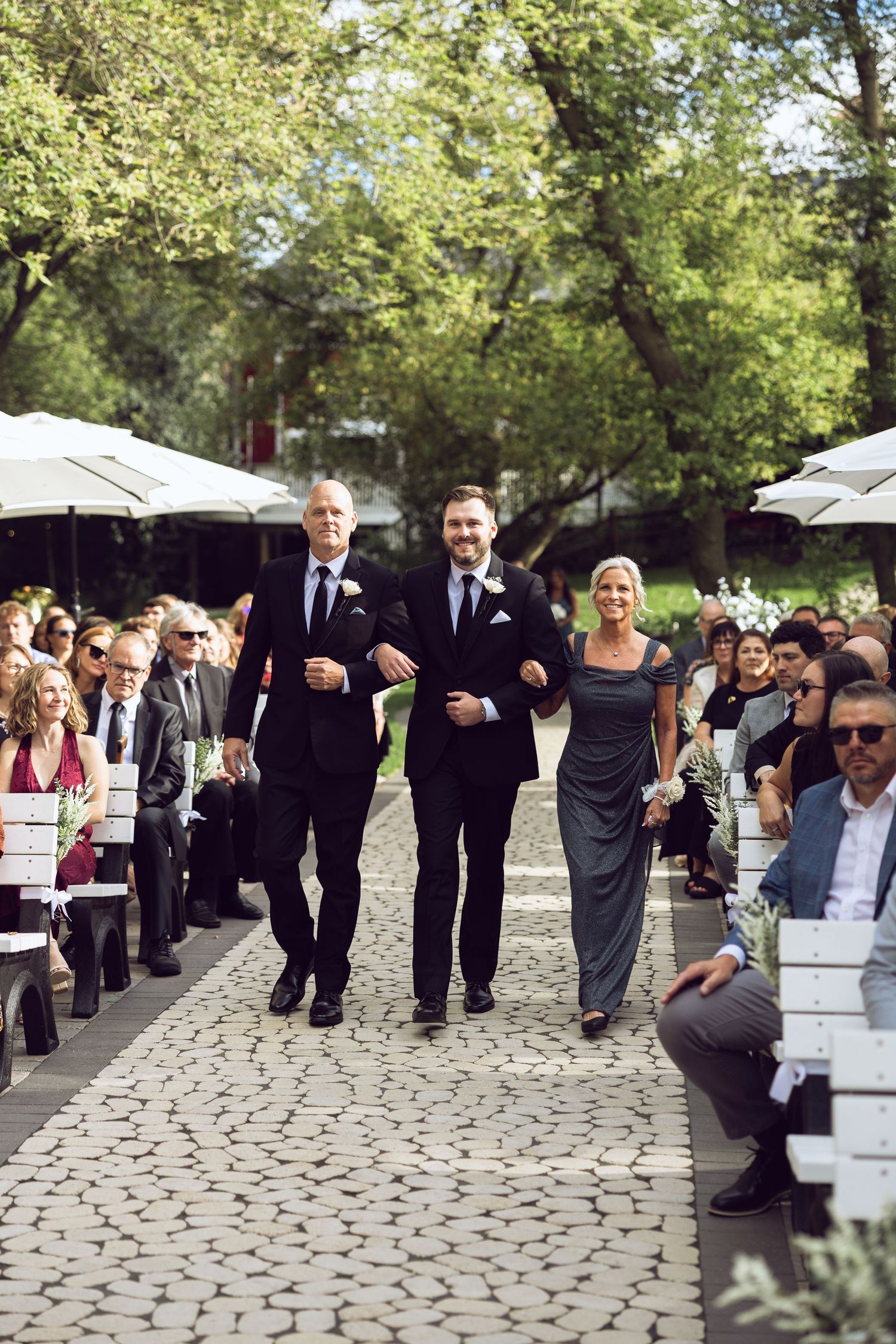 Groom walks down a cobblestone aisle between seated guests, escorted by two men in suits and a woman in a gray gown.