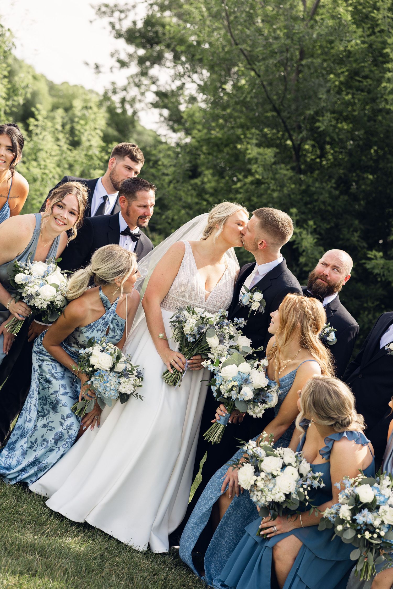 Bride and groom share a kiss surrounded by bridesmaids in blue dresses holding white bouquets and groomsmen on a grassy lawn.