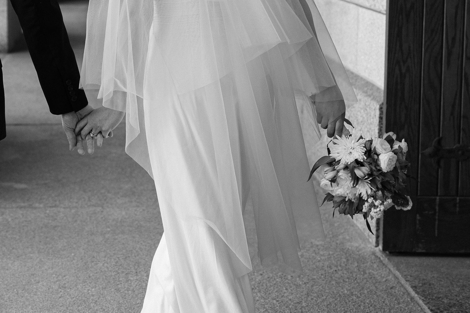 A black and white close-up of a bride in a veil holding her groom's hand and a bouquet of flowers as they walk together.