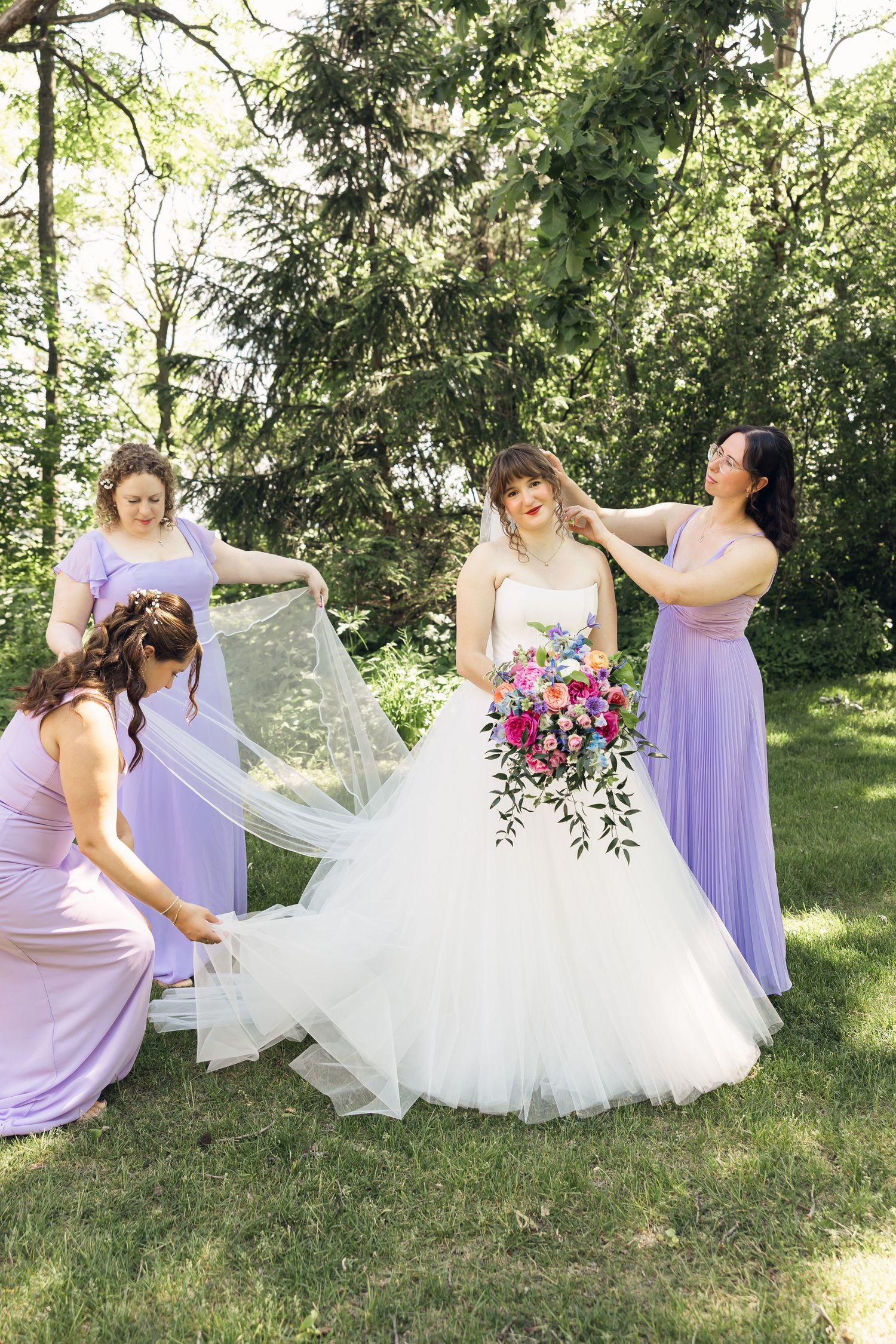Bridesmaids in lavender dresses adjust the bride's veil and hair on the lawn as she holds a vibrant pink floral bouquet.