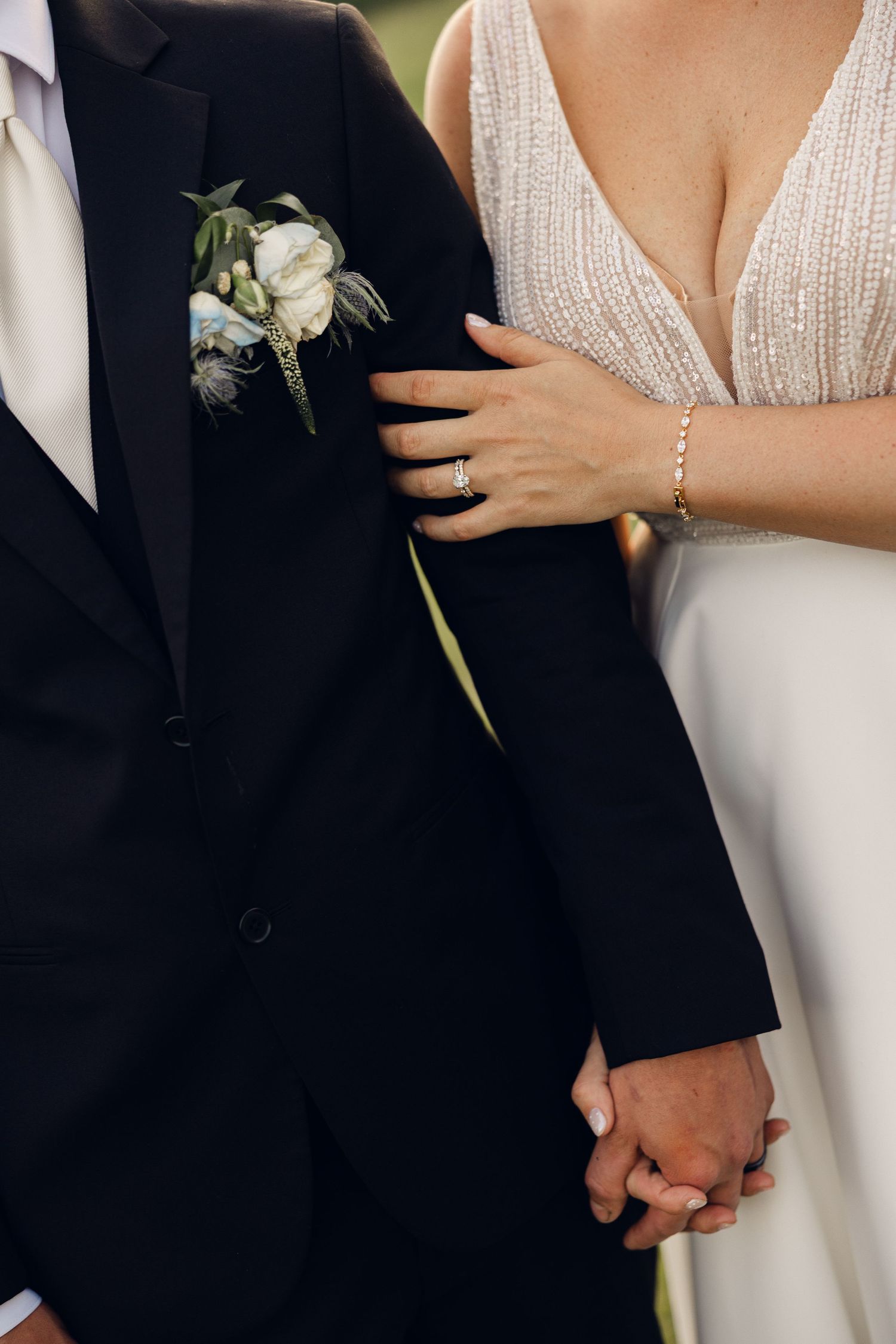 Close-up of a bride and groom holding hands, with the groom's white floral boutonniere and the bride's sparkling ring visible.