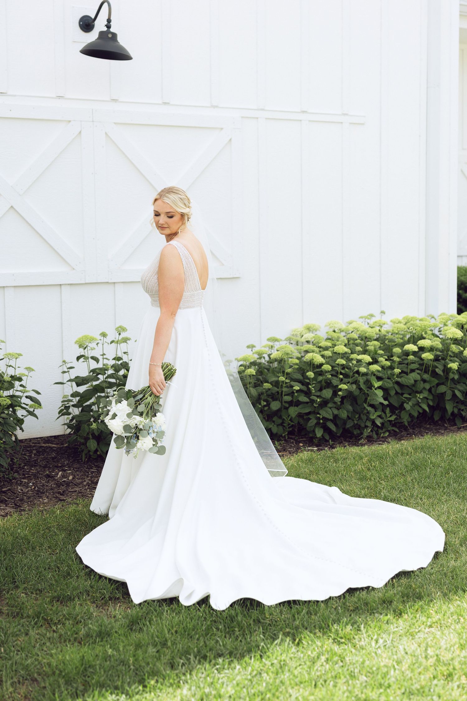 A bride in a flowing white gown with a long train holds a white floral bouquet while standing on grass beside a white barn.