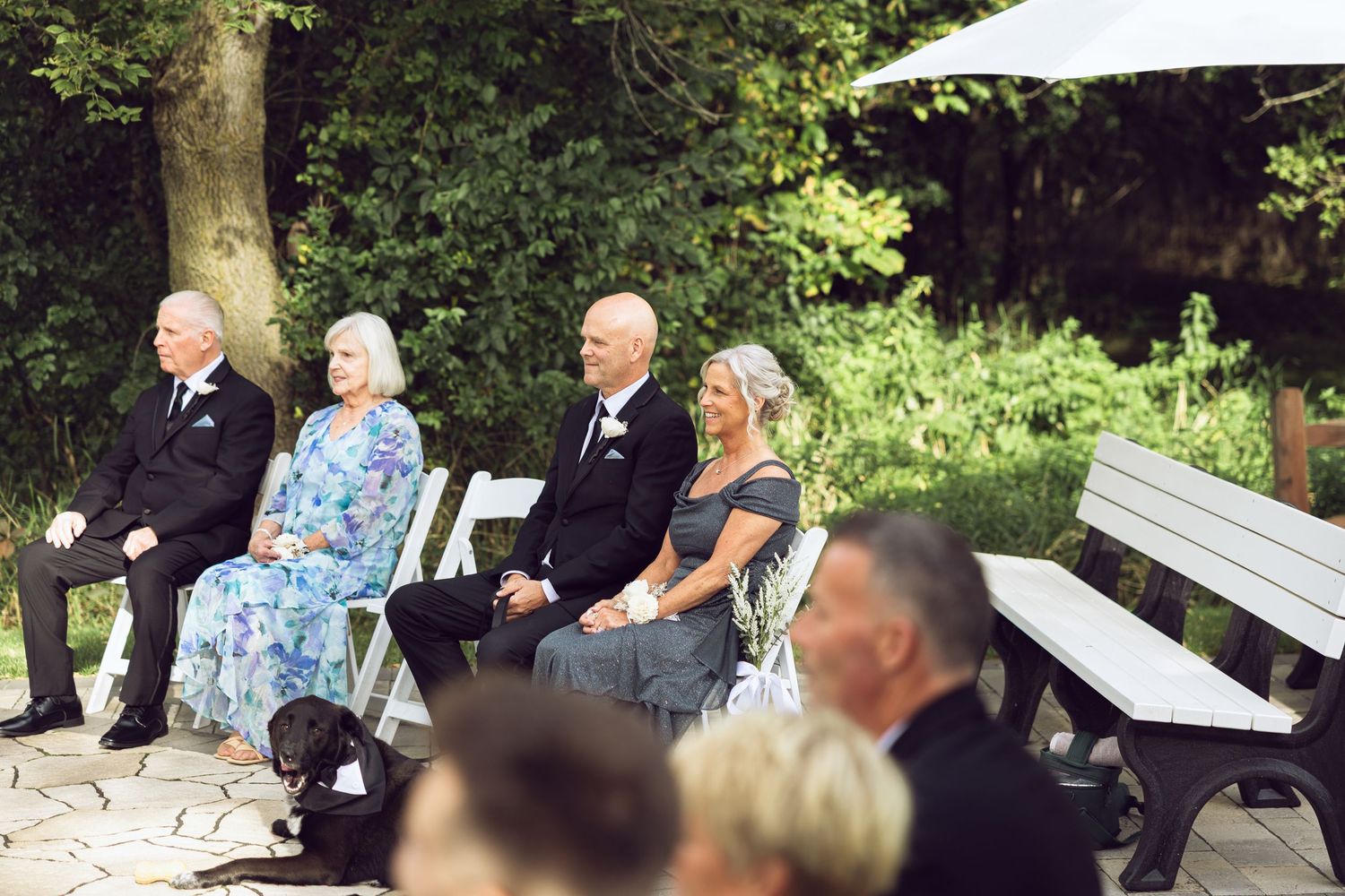 Seated wedding guests in suits and dresses watch an outdoor ceremony while a small black dog rests on the stone patio.
