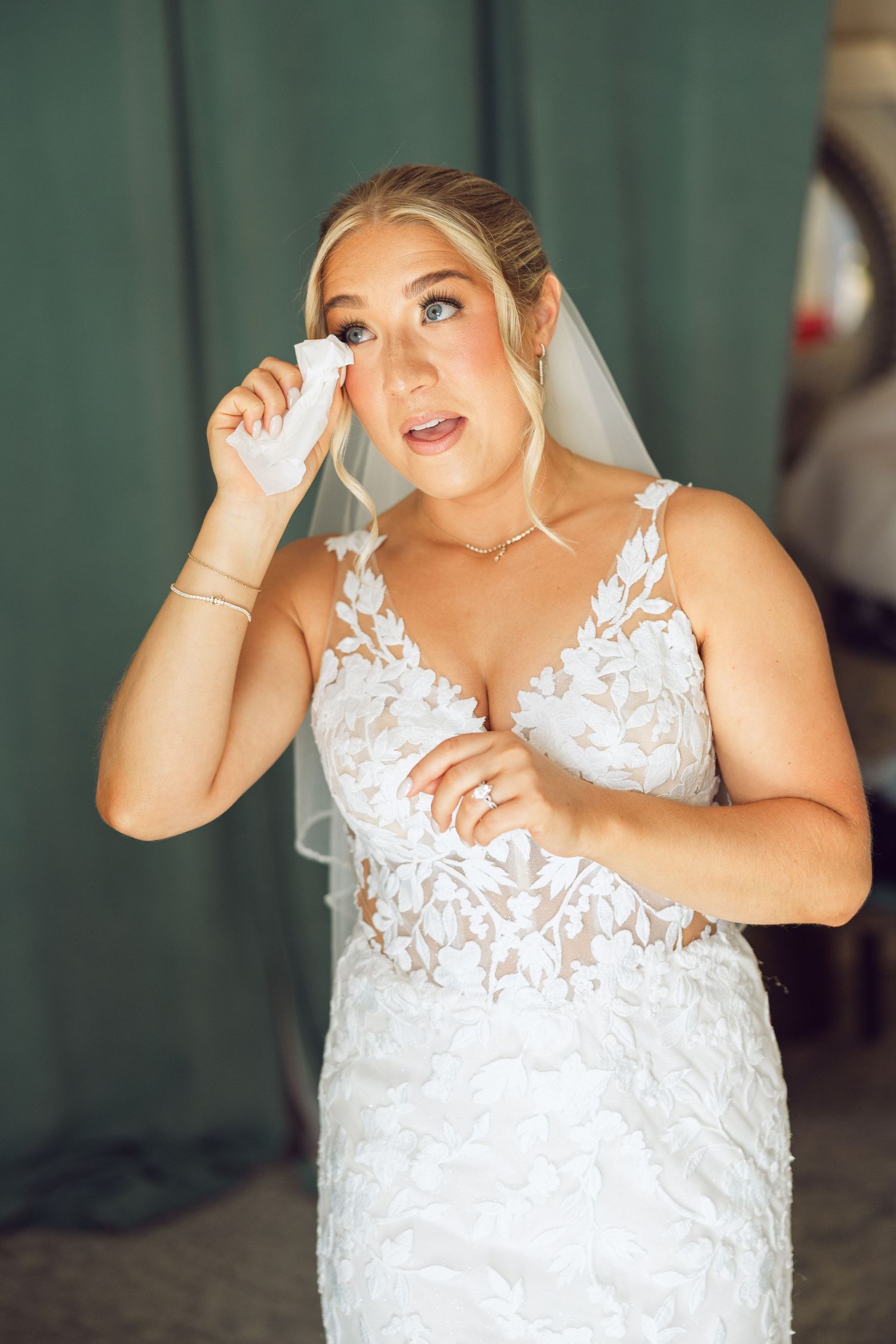 Bride in a lace wedding gown and veil dabs a tear with a tissue while getting ready on her wedding day.