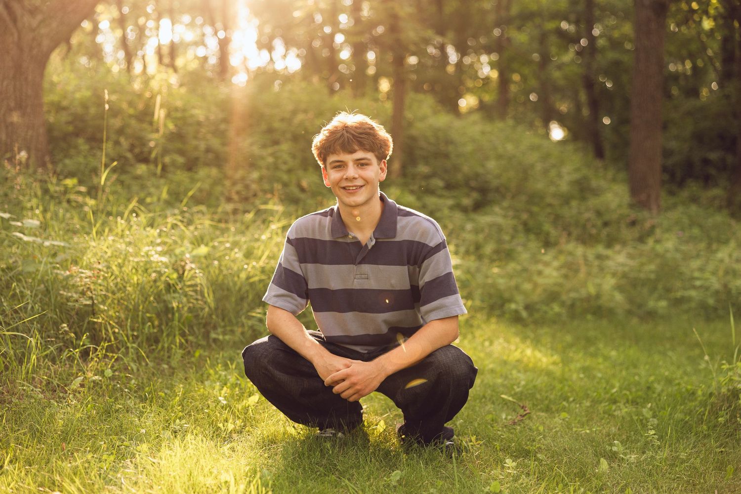 Smiling teenage boy in a striped shirt crouches on a grassy lawn with warm sunlight filtering through the trees behind him.