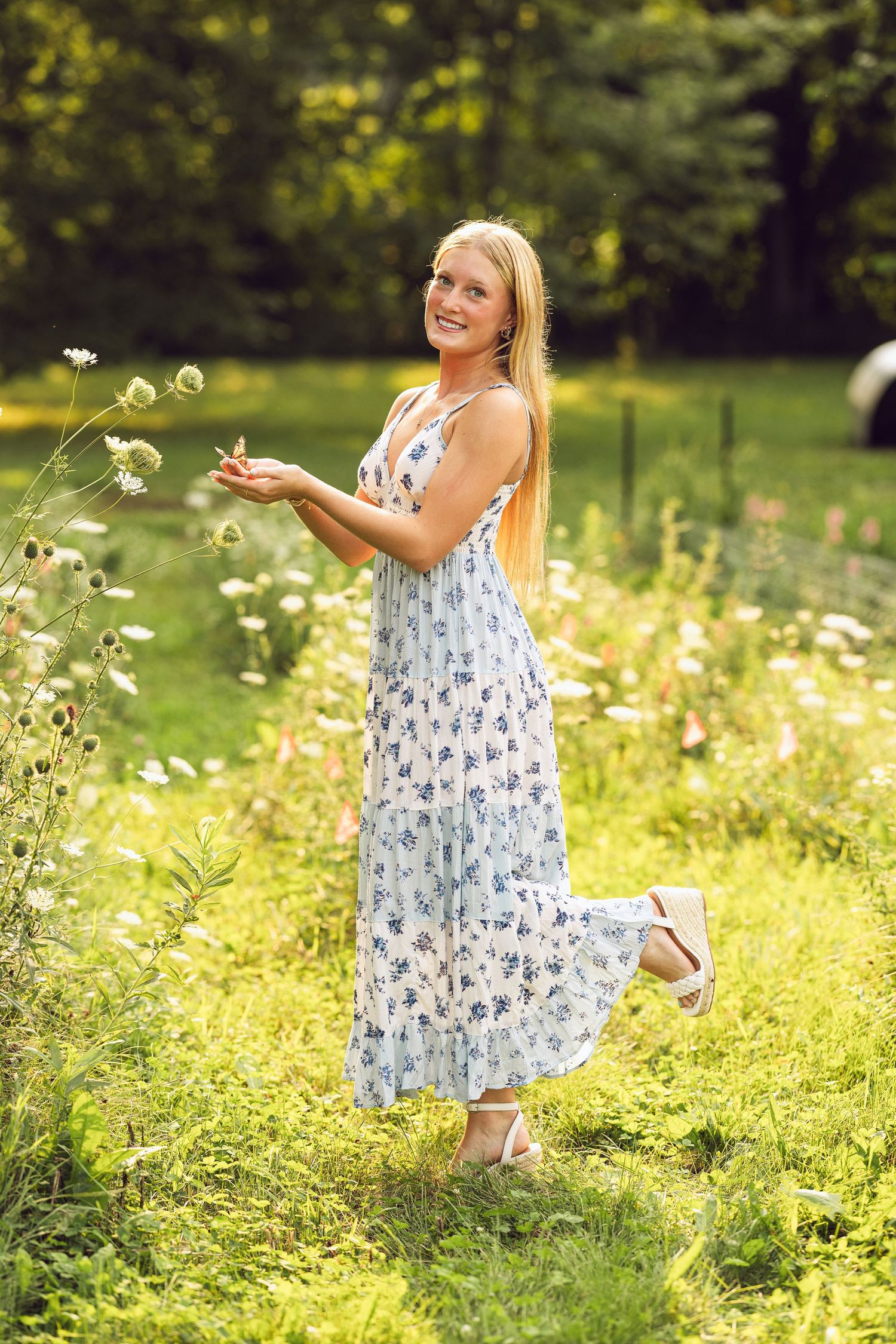Smiling young woman with long blonde hair in a floral blue maxi dress stands in a sunny field of wildflowers.