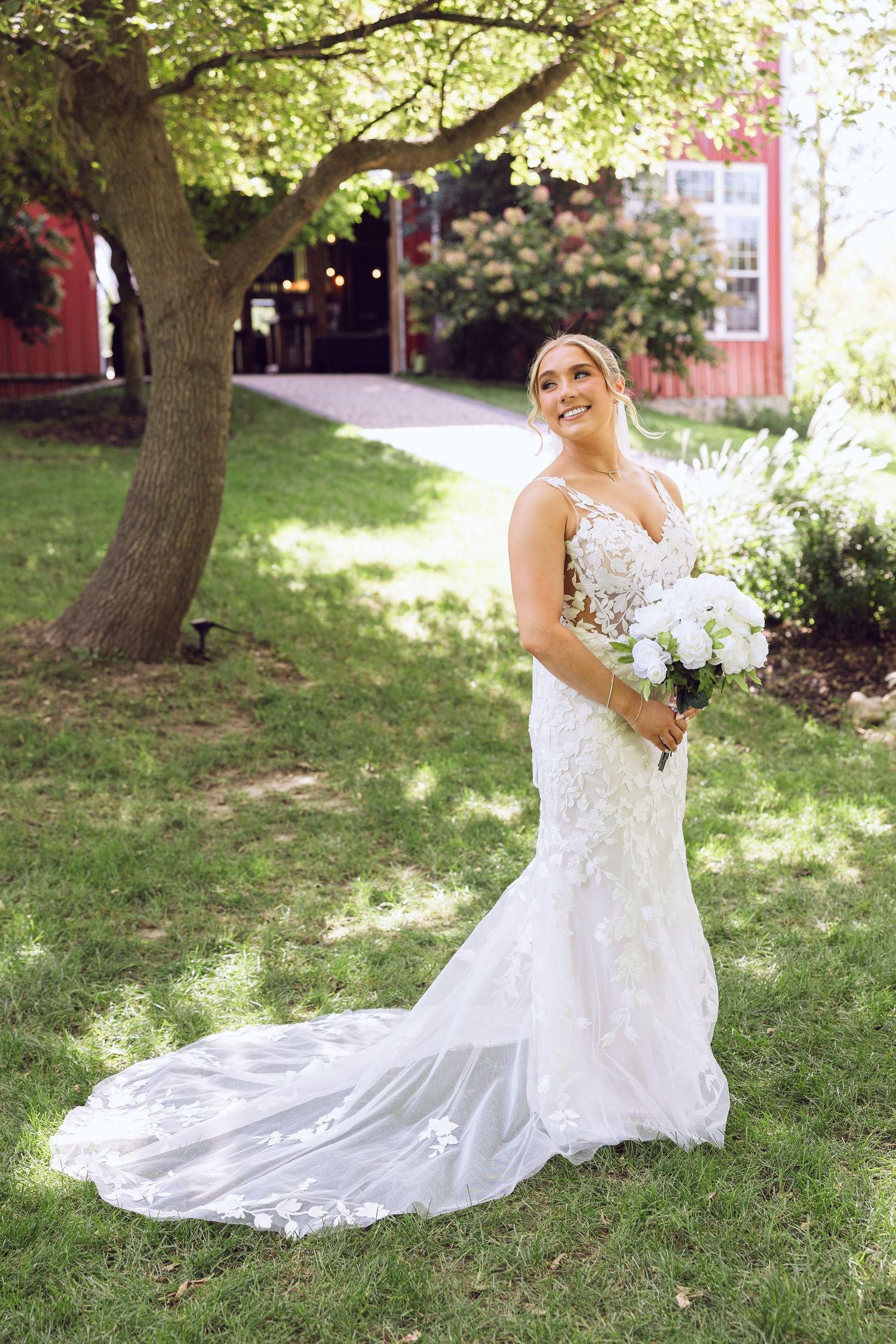 Smiling bride in a lace mermaid wedding dress with a long train holds a white bouquet on a green lawn near a red barn.