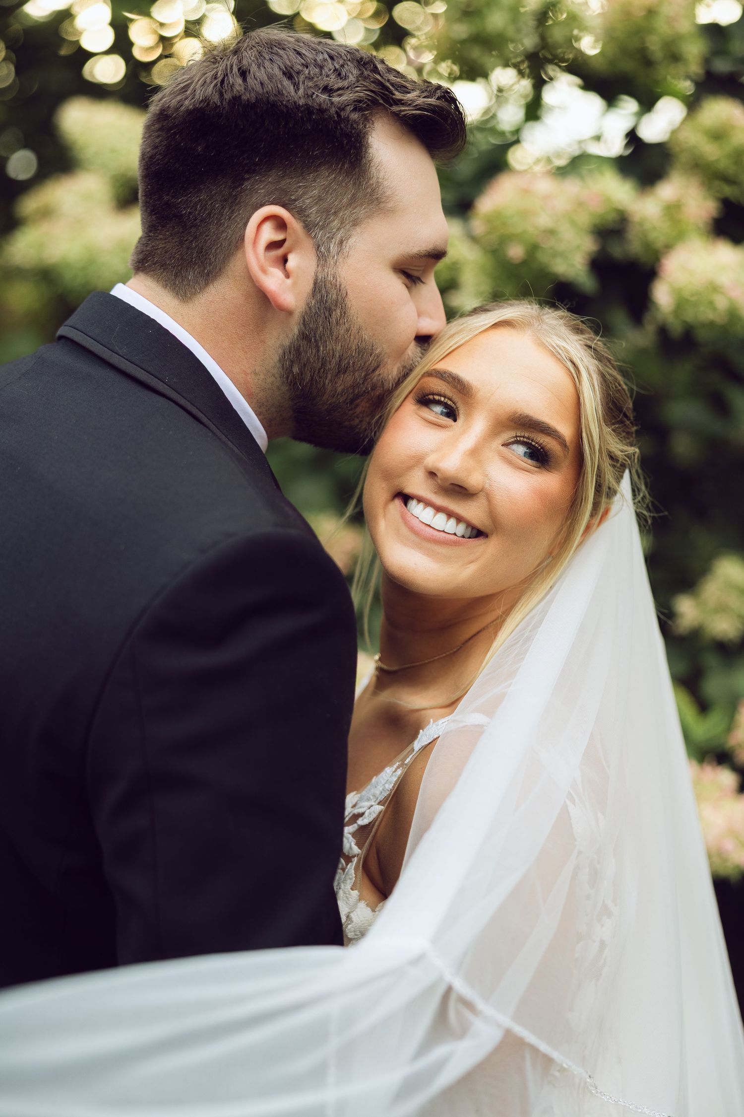 A bearded groom kisses the cheek of his smiling bride in a flowing veil in front of blooming hydrangeas.