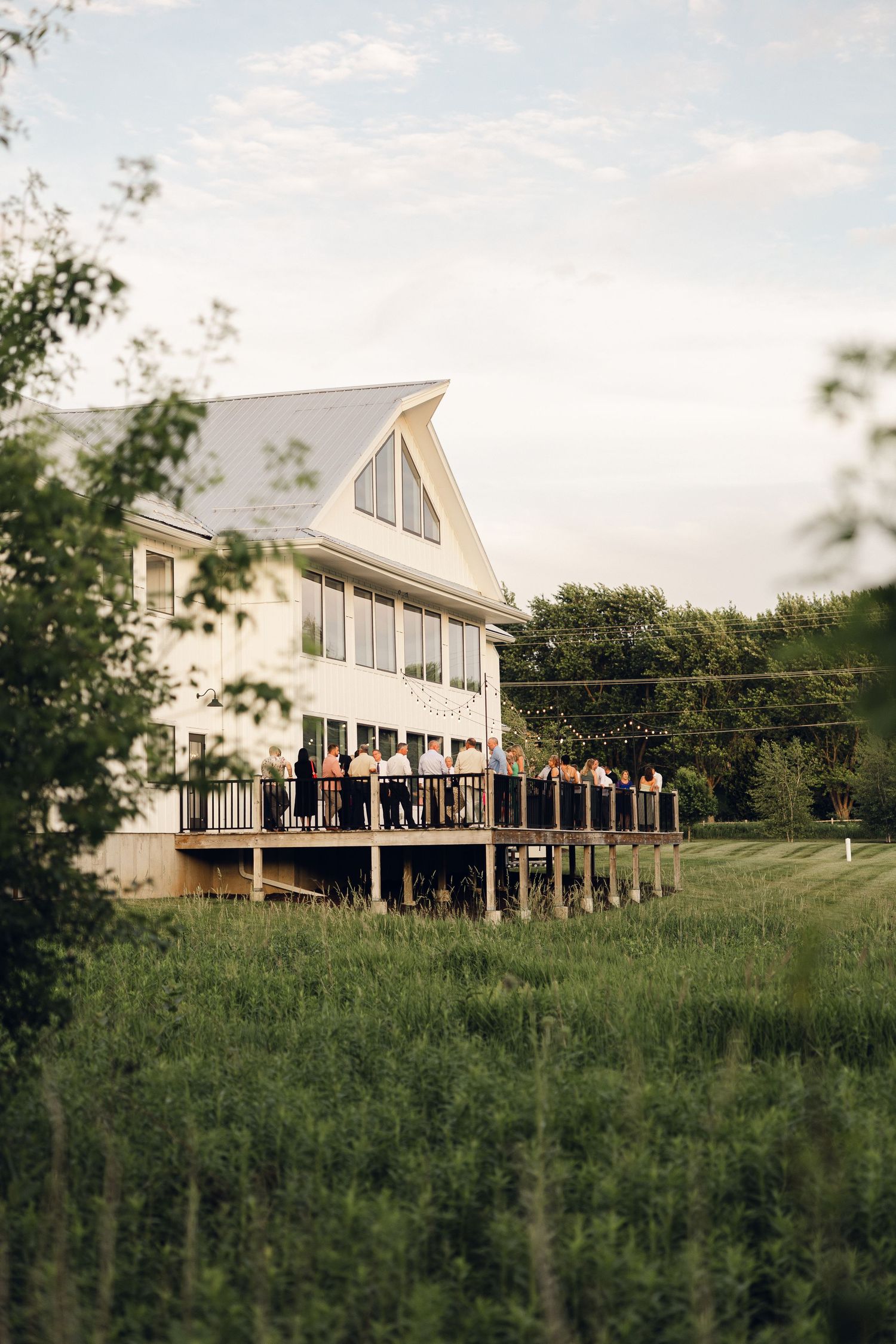 White venue with a large wraparound deck where wedding guests gather, set among green grass and trees under a soft evening sky.