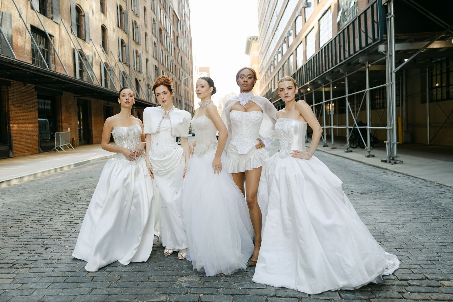 Five models in elegant white bridal gowns pose together on a cobblestone city street lined with brick buildings.