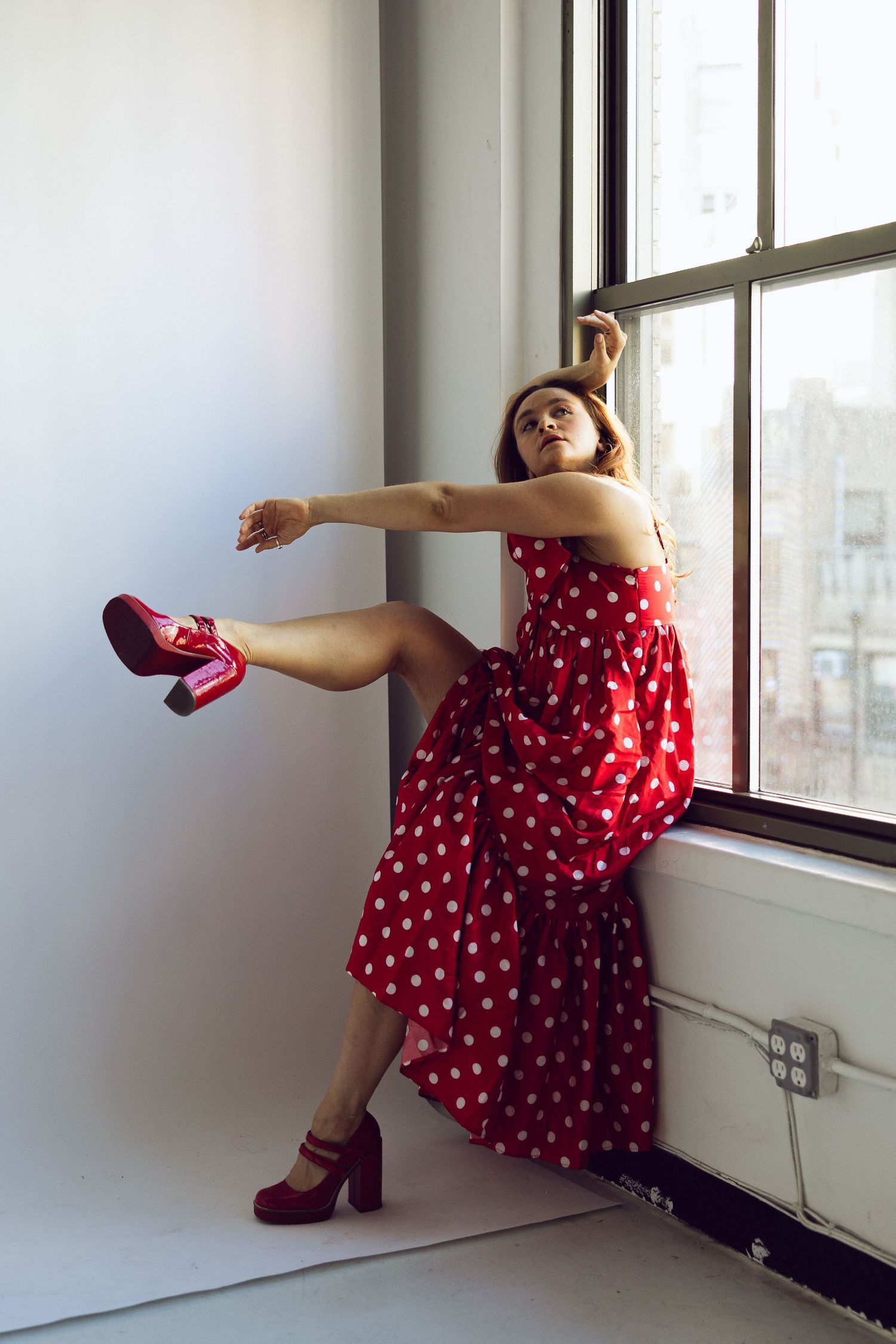 Woman in a red polka dot dress and platform heels sits on a window ledge in a bright loft, one leg stretched out.
