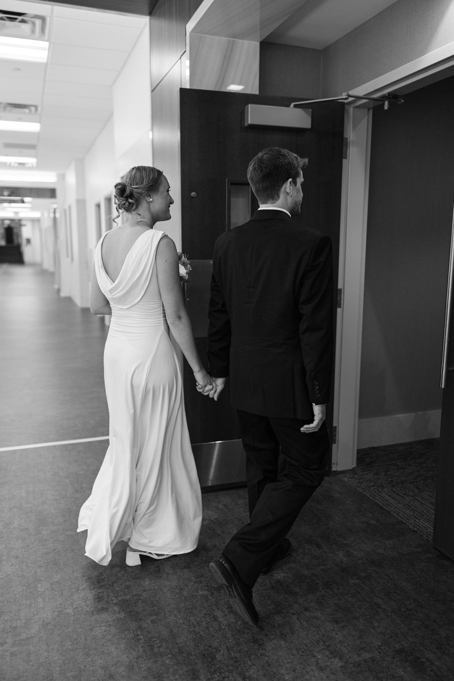 Bride in a white gown and groom in a dark suit hold hands as they walk down a hallway in this black and white wedding photo.