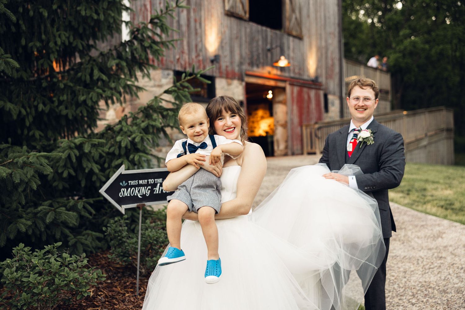Bride and groom smile with a toddler ring bearer near a rustic barn venue and a wooden dancing arrow sign.