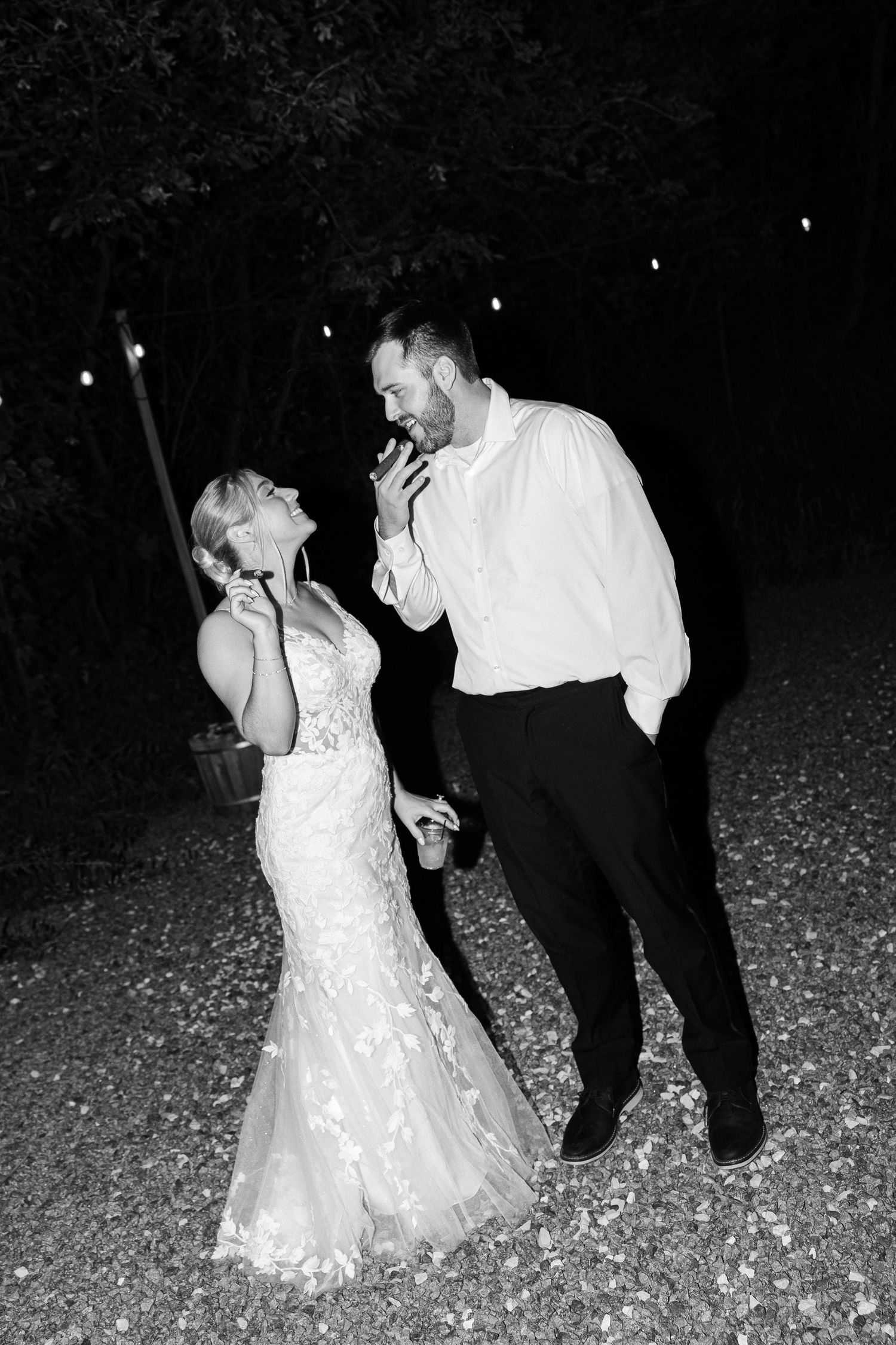 A black and white photo shows a bride and groom laughing together outdoors at night on a petal-covered path under string lights.