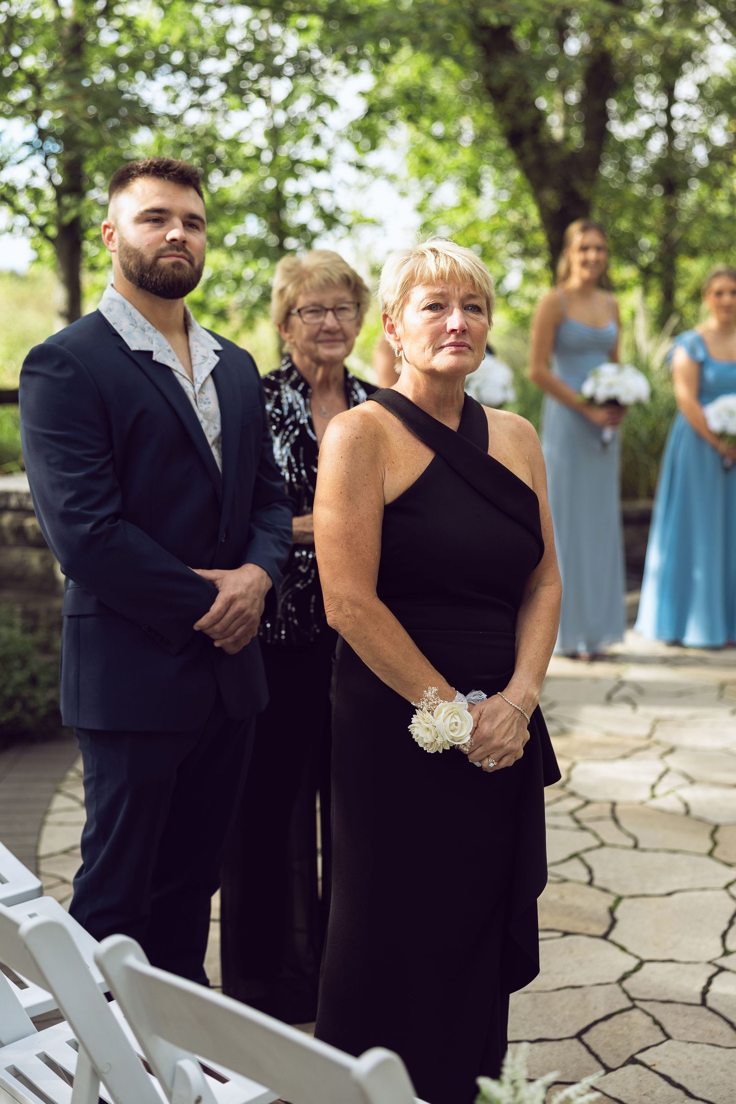 Wedding guests in formal attire stand during an outdoor ceremony, including a woman in a black dress wearing a flower wrist corsage.