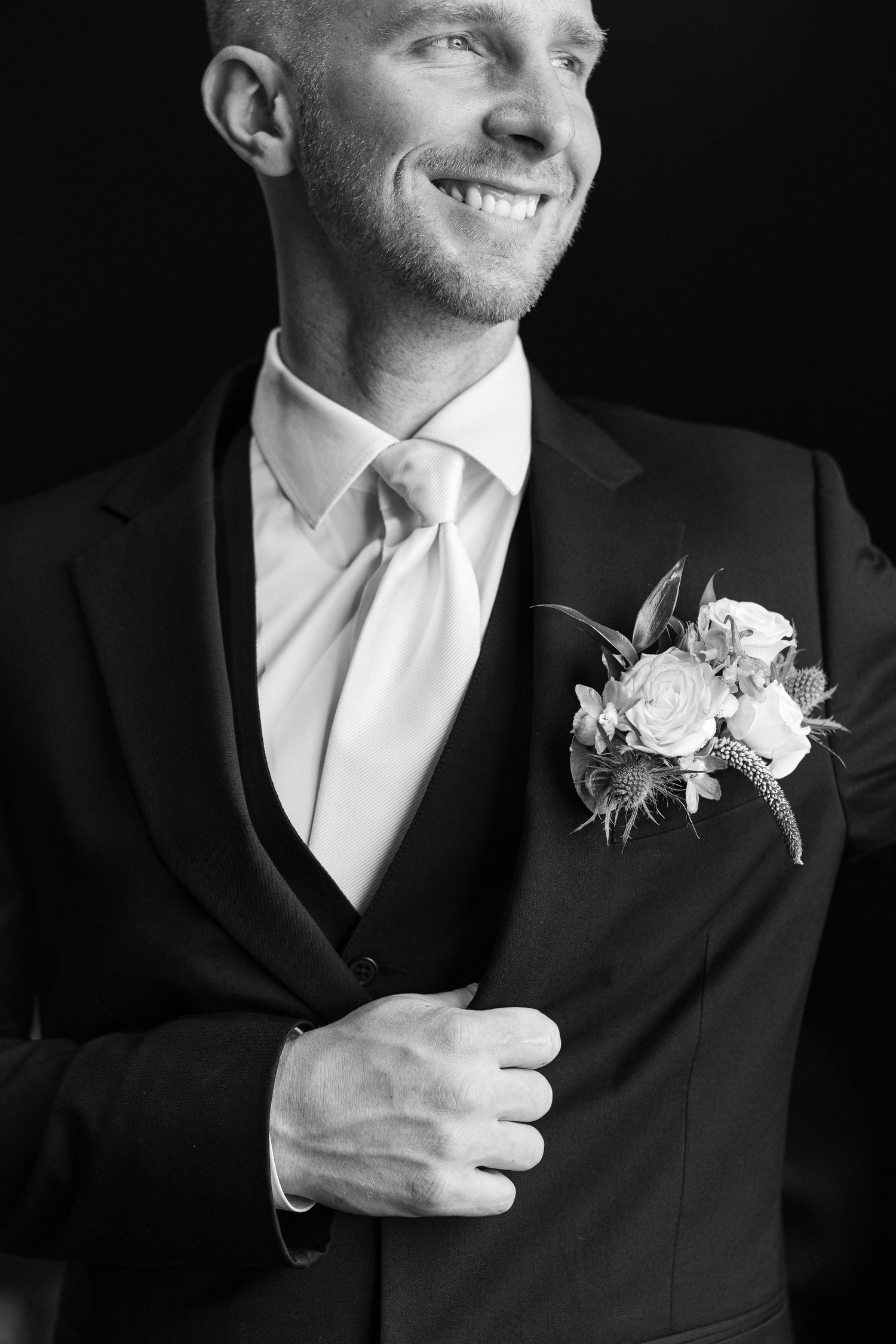A smiling groom in a dark suit, vest, and white tie adjusts his jacket, wearing a rose boutonniere, in a black and white portrait.