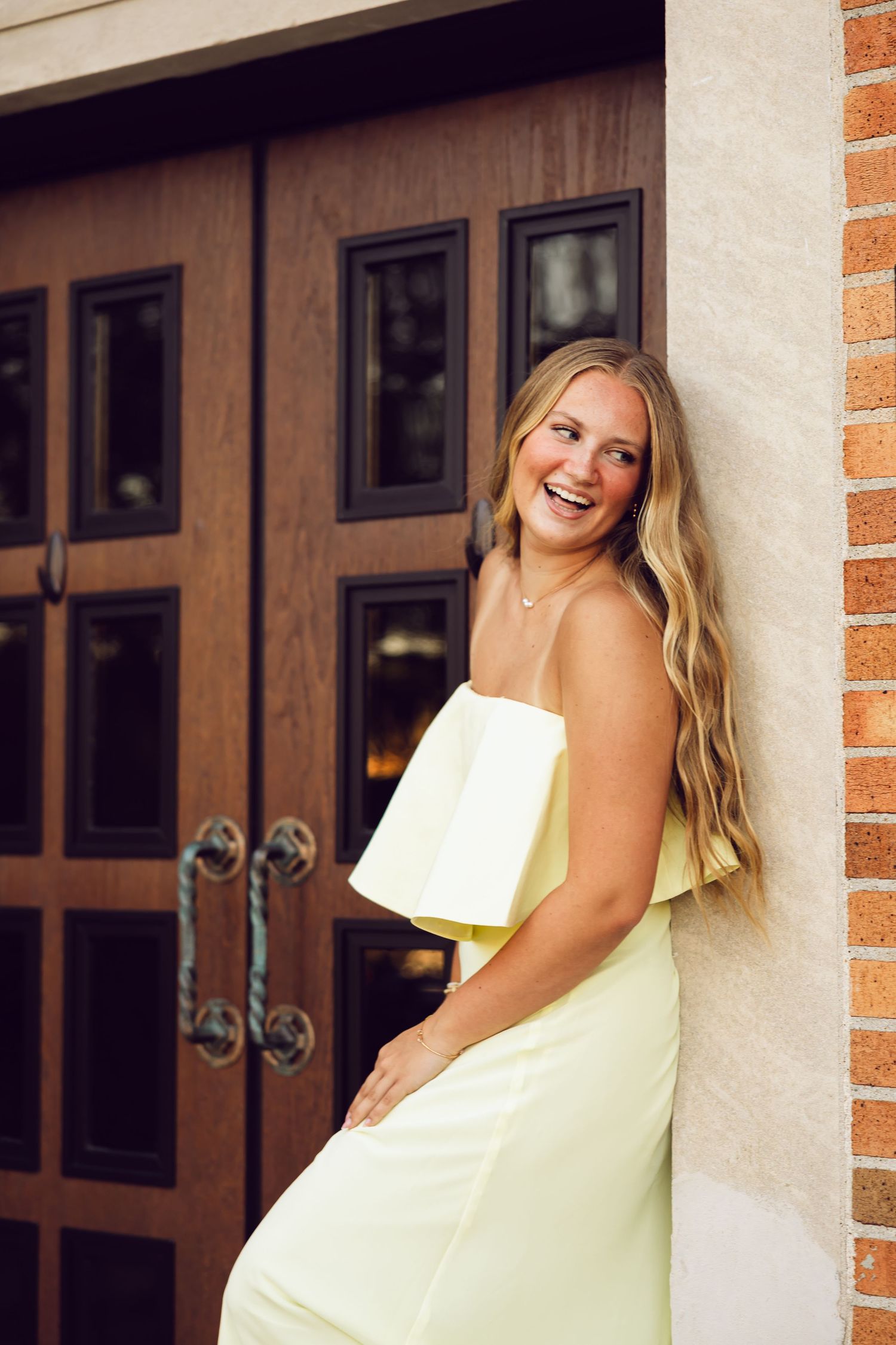 Laughing young woman in a pale yellow strapless dress leans against a brick wall beside large wooden doors.