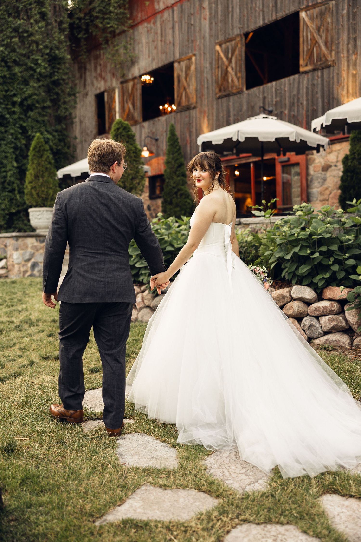 Bride and groom walk hand in hand along stone steps toward a rustic wooden barn wedding venue with string lights.