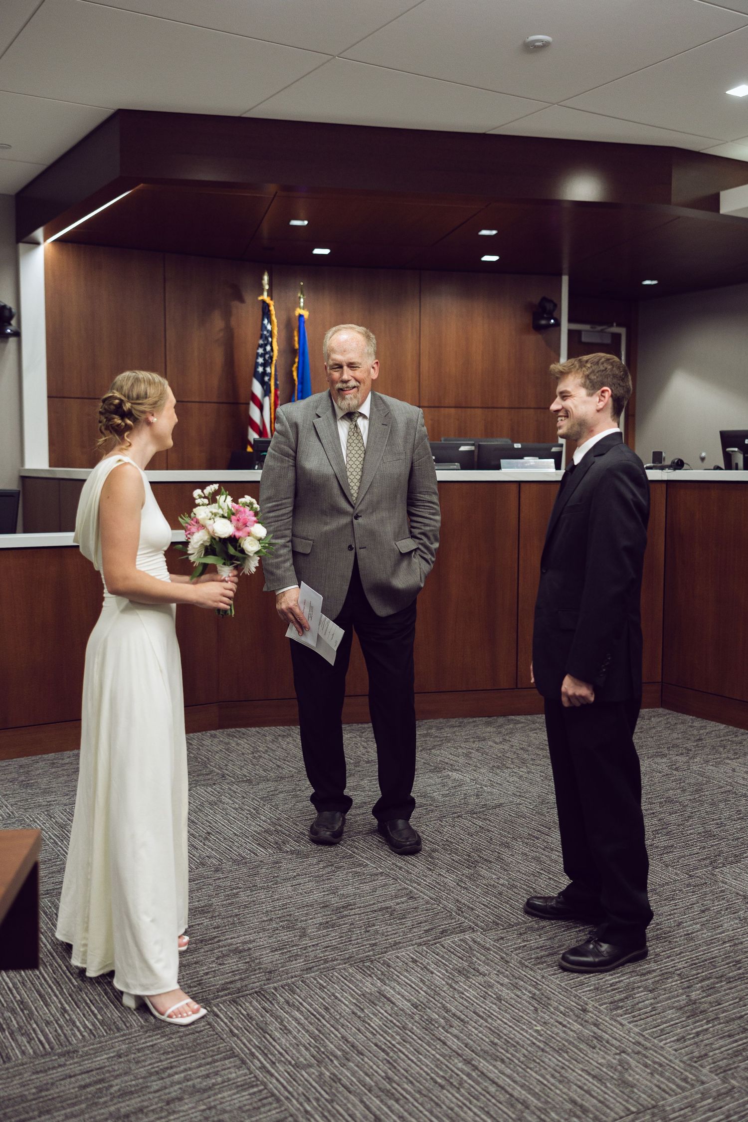 Bride holding a bouquet and groom in a black suit face a smiling officiant during a courthouse wedding ceremony.