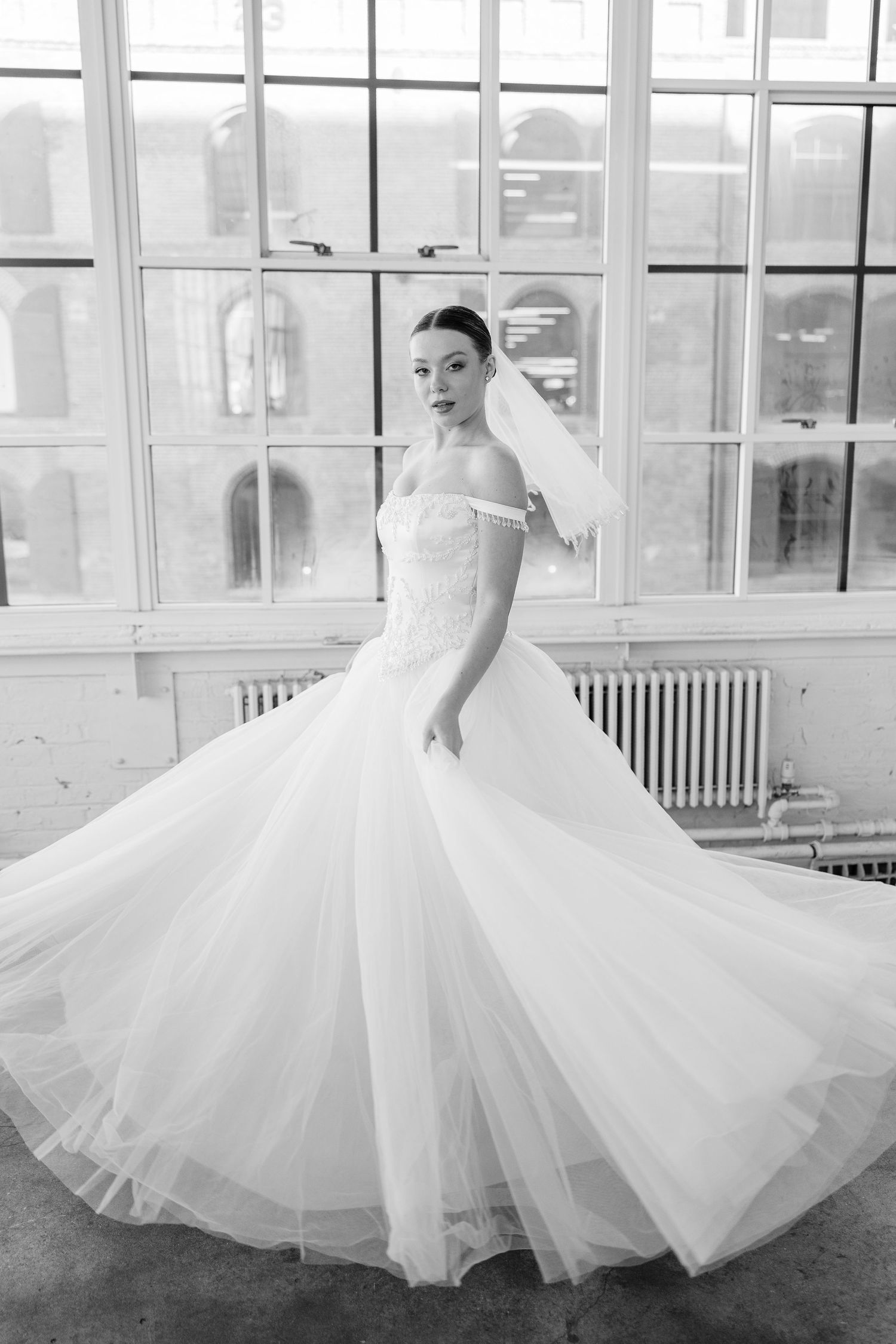 Black and white image of a bride in a flowing tulle ball gown and short veil spinning in a window-lit room.