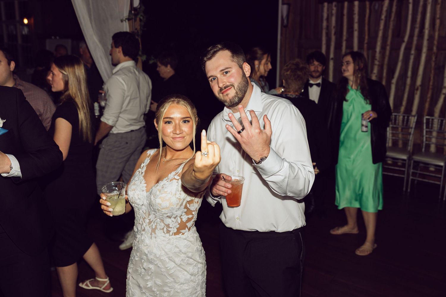 A smiling bride in a lace gown and groom in a white shirt pose with drinks among guests at a dimly lit evening reception.