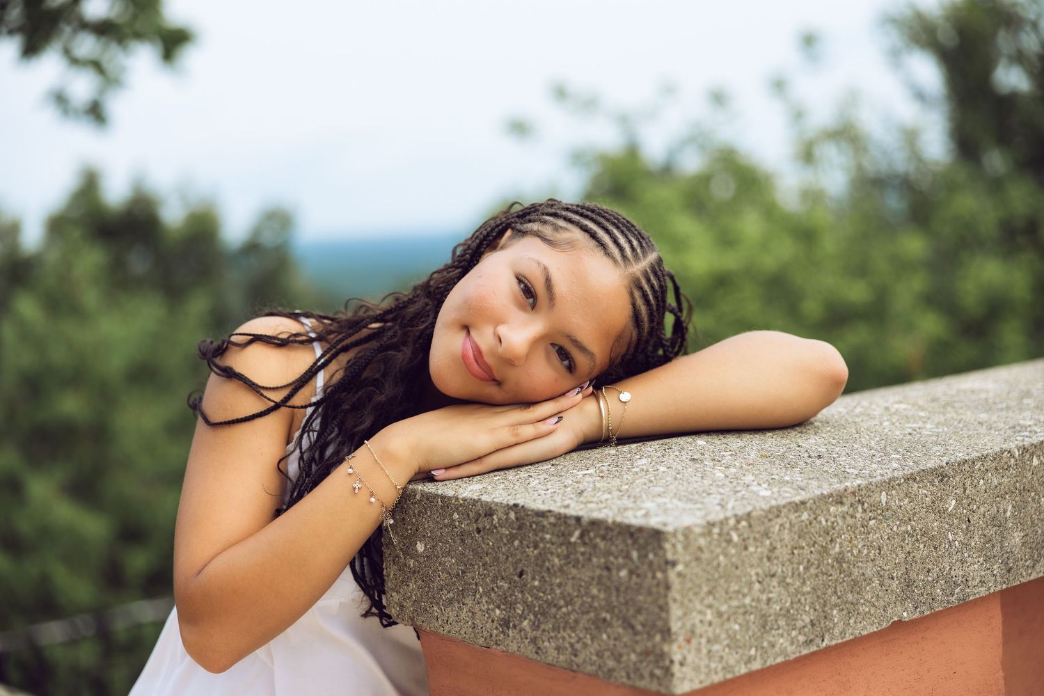 Smiling young woman with long braided hair rests her head on her folded arms on a stone ledge with green trees behind.