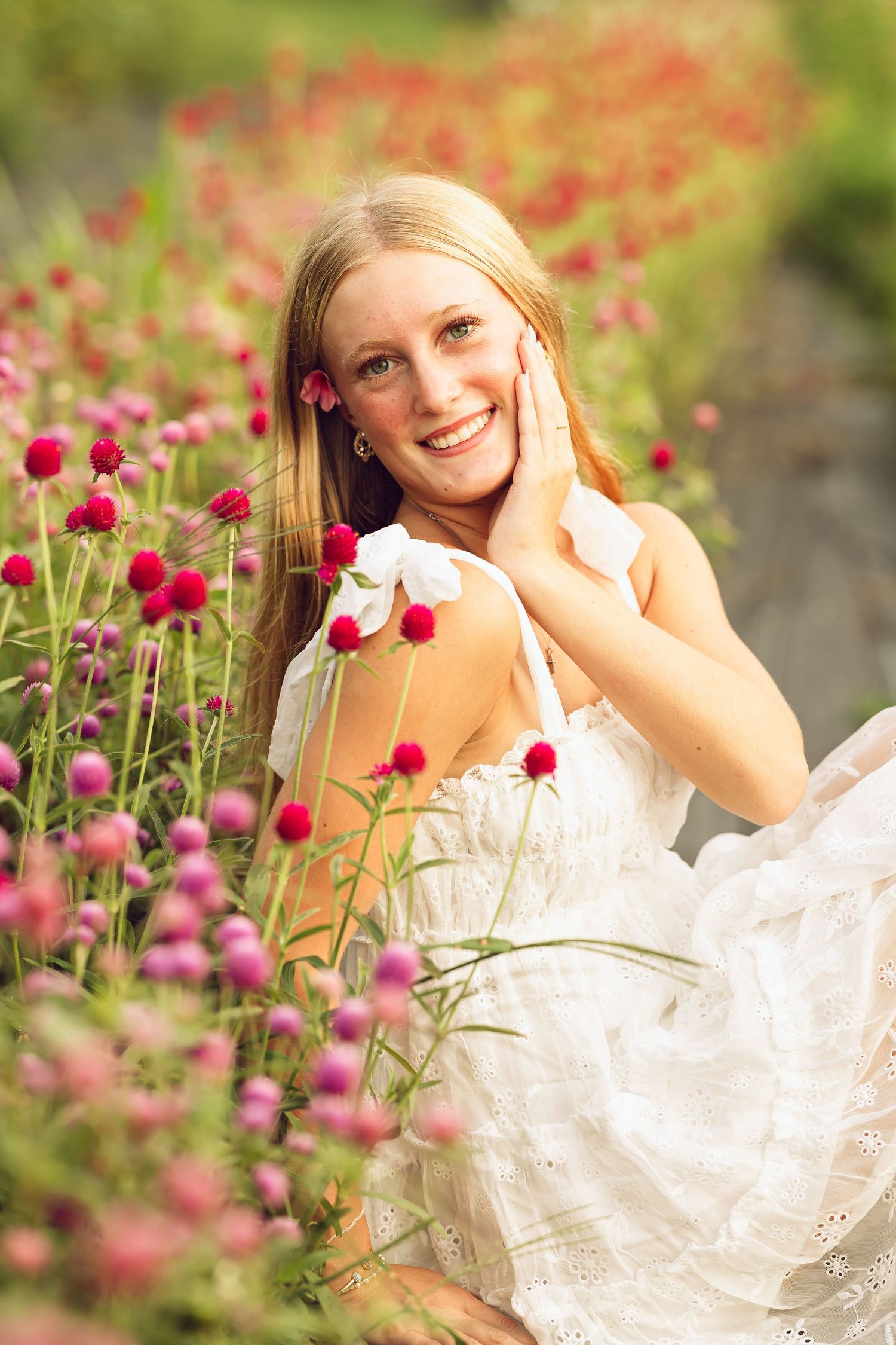 Smiling blonde young woman in a white dress rests her cheek on her hand among pink and red globe amaranth flowers.