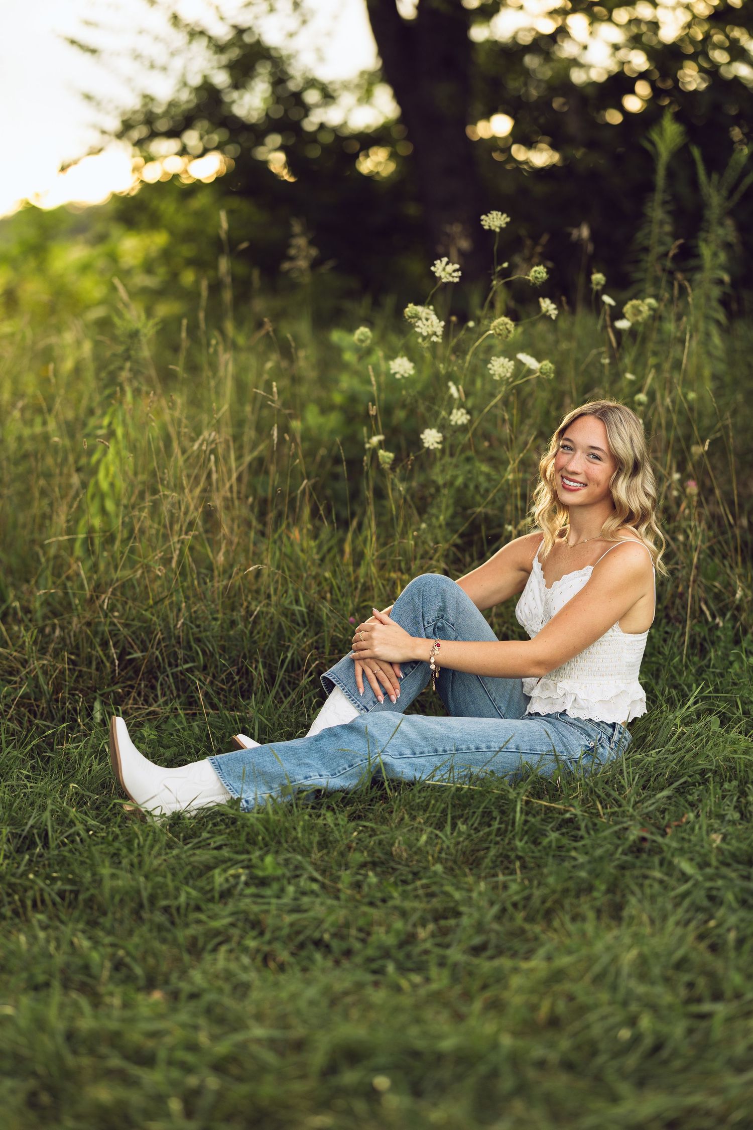 Smiling young woman with wavy blonde hair sits in a sunlit grassy field wearing a white top, light-wash jeans and white boots.