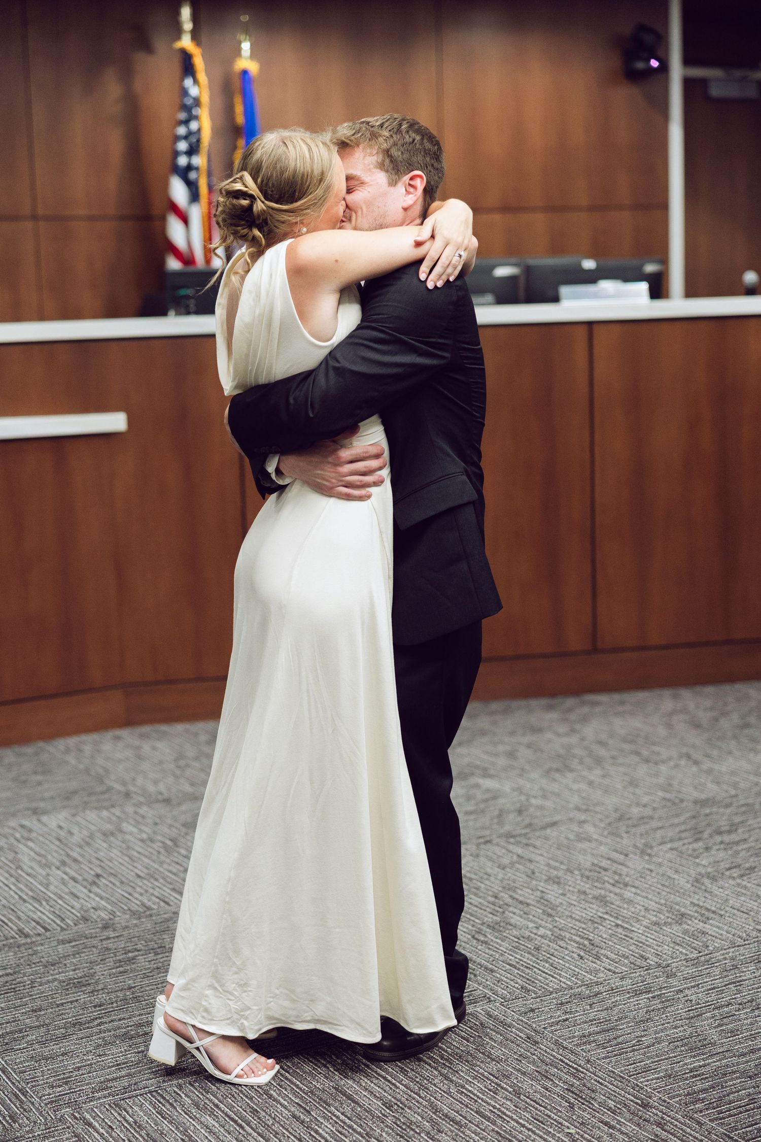Newlywed couple share a tender kiss and embrace in a courthouse, the bride in a white gown and the groom in a black suit.