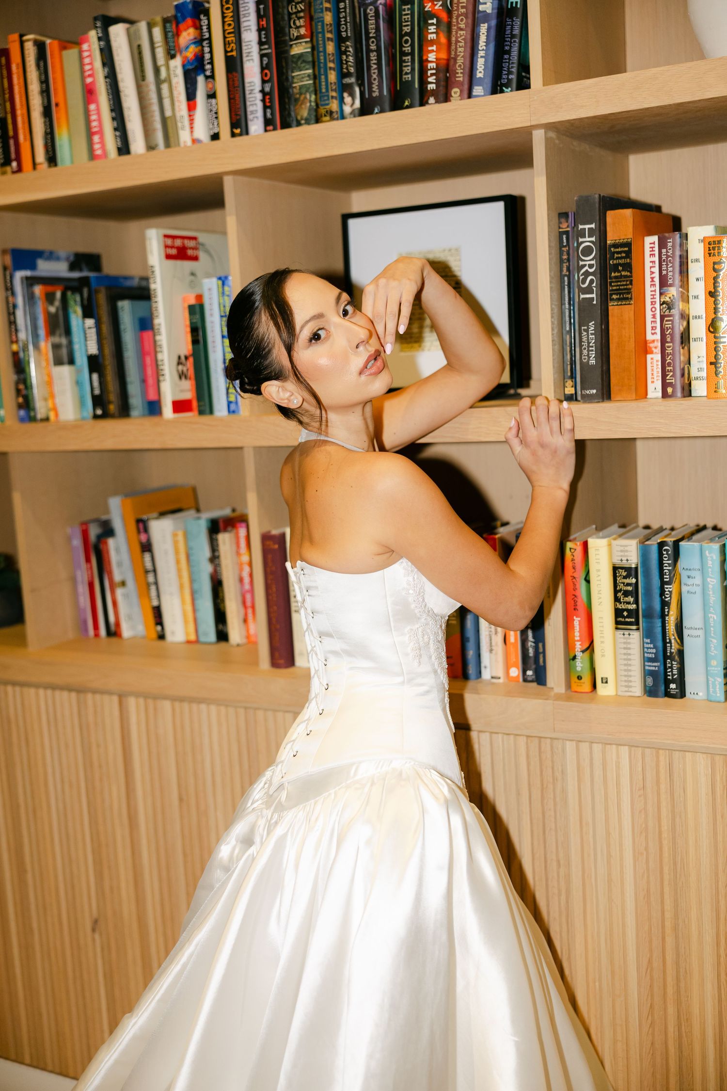 Bride in a strapless satin corset-back wedding gown poses with raised arms in front of colorful bookshelves.