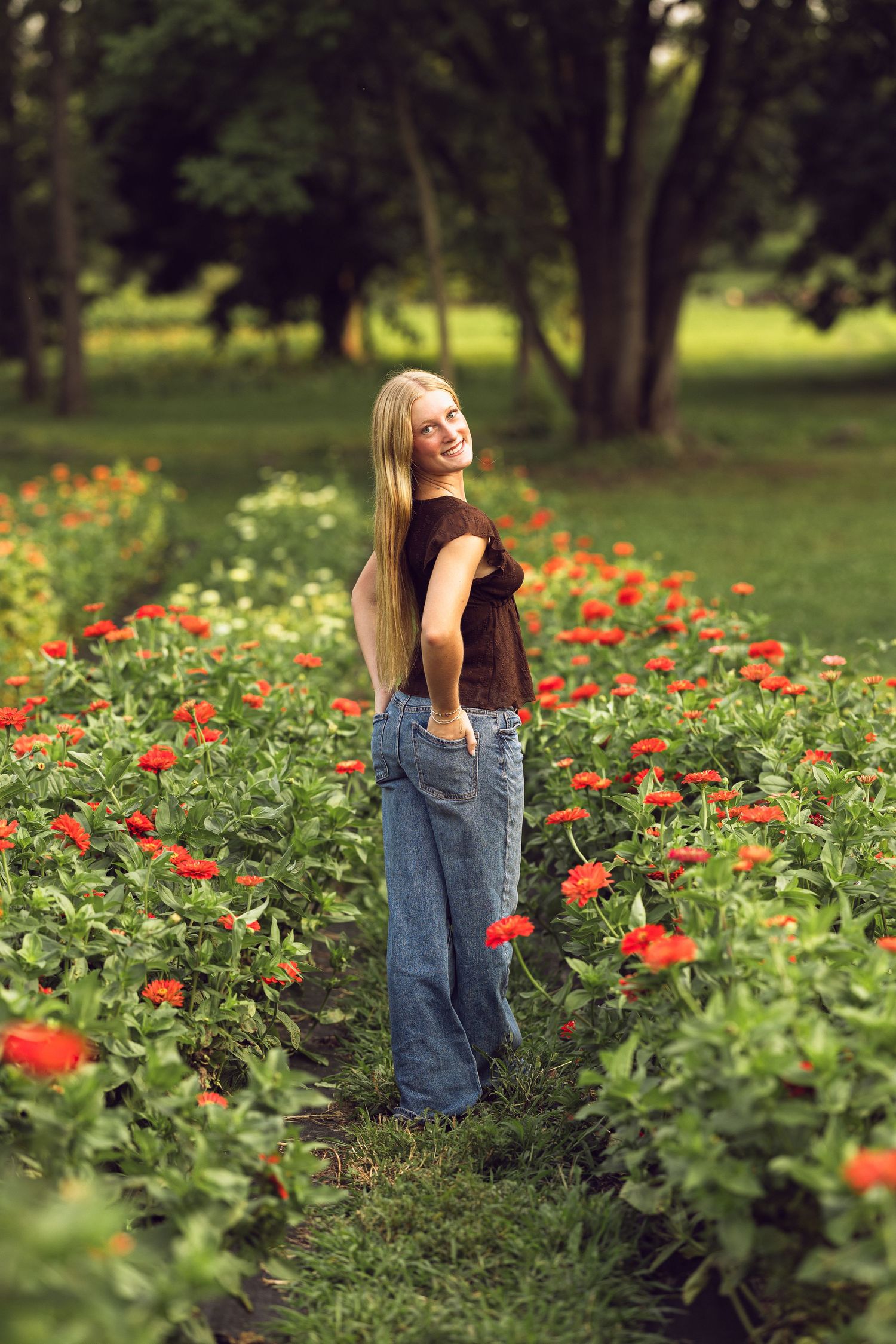 Smiling blonde young woman in jeans looks over her shoulder while standing on a path among bright orange zinnia flowers.
