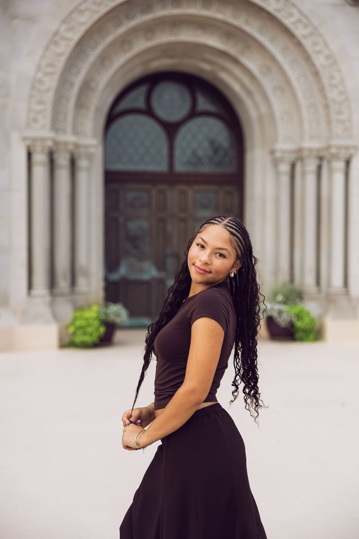 Young woman with long braids and a dark outfit smiles over her shoulder in front of an ornate arched stone doorway.