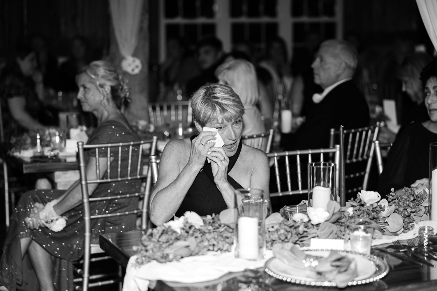 In a black and white photo, a woman at a reception table wipes her tears with a napkin while other guests sit nearby.