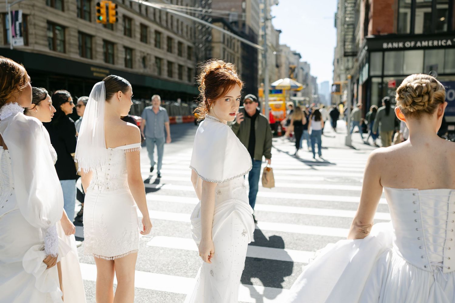 Four models in white bridal gowns and veils stand together on a busy city crosswalk with tall buildings behind them.