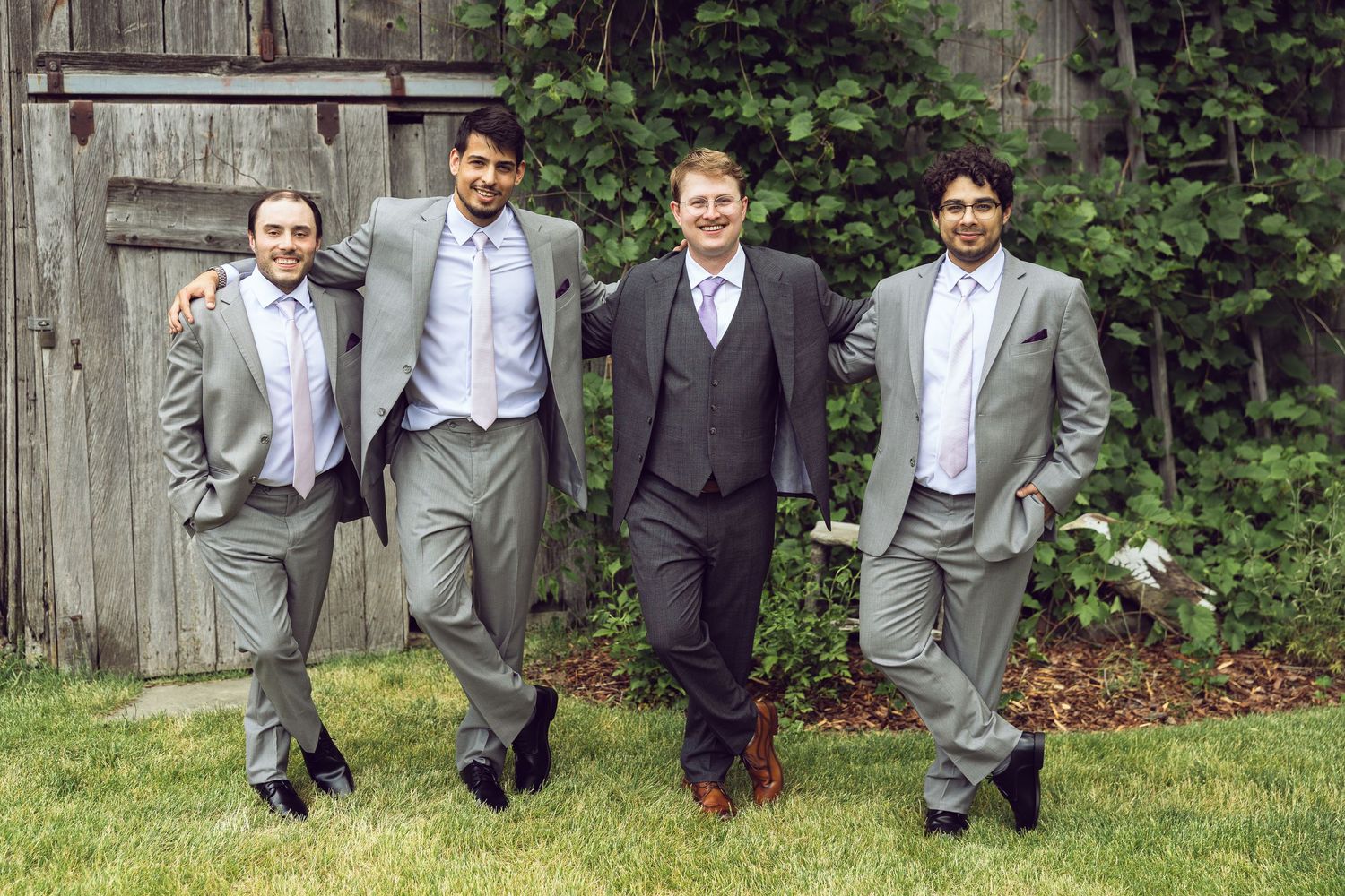 Groom and three groomsmen in gray suits pose playfully on a grassy lawn in front of a rustic barn and green ivy.