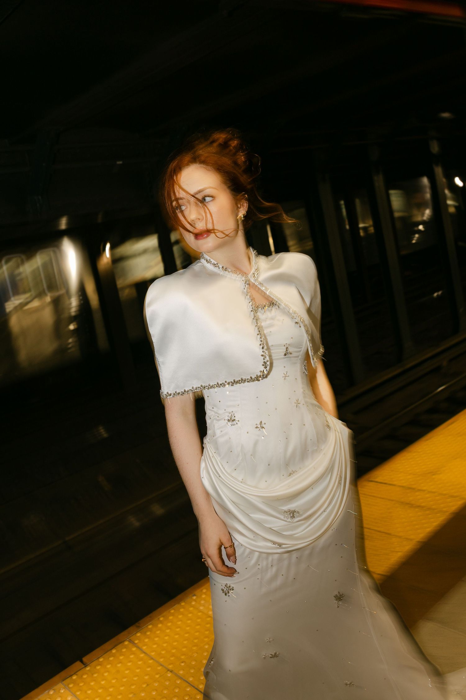 Bride with auburn hair in a beaded white dress and satin capelet glances sideways on a yellow-edged subway platform.
