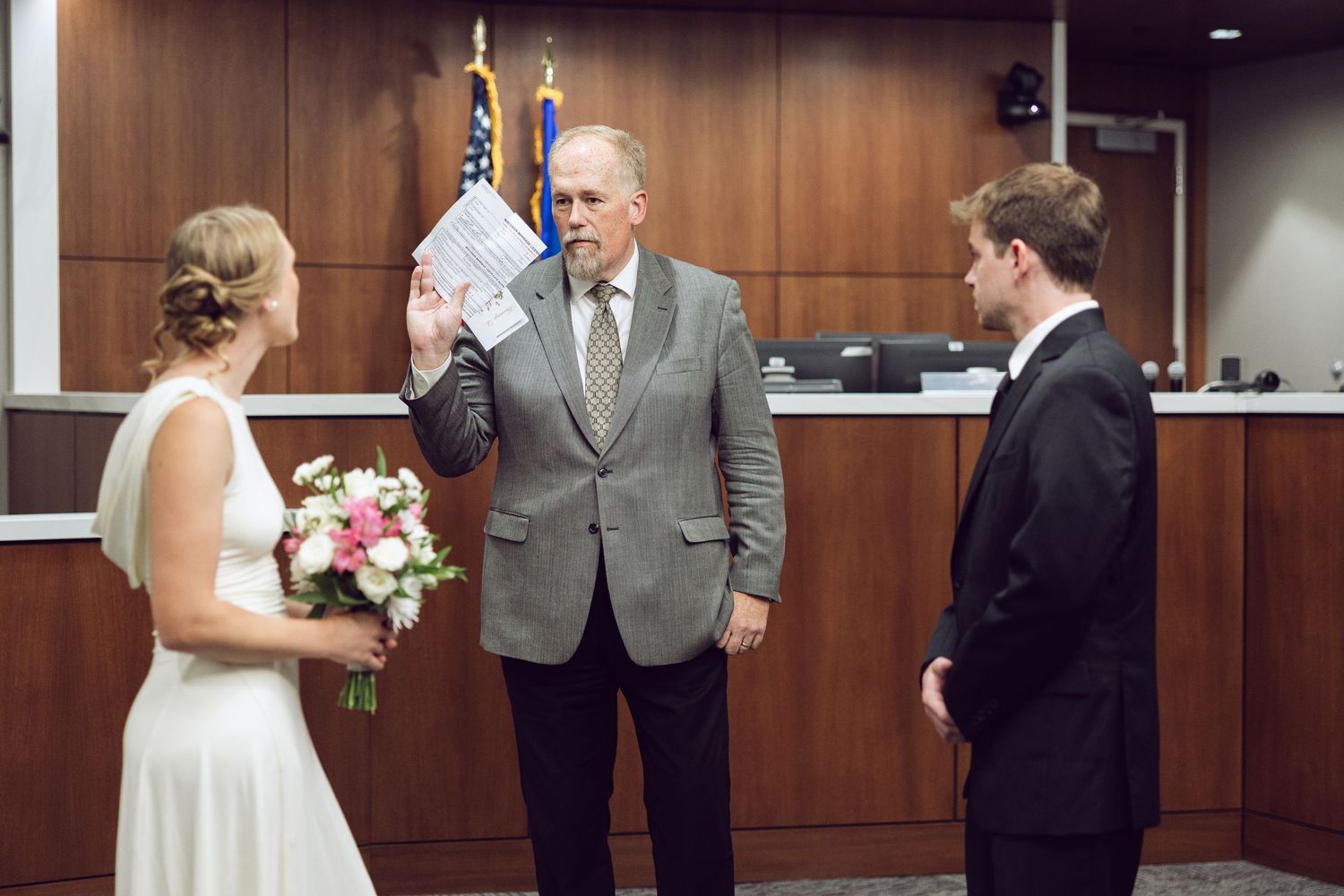 Officiant raises his right hand while holding a paper as the bride and groom stand before him during their courthouse vows.