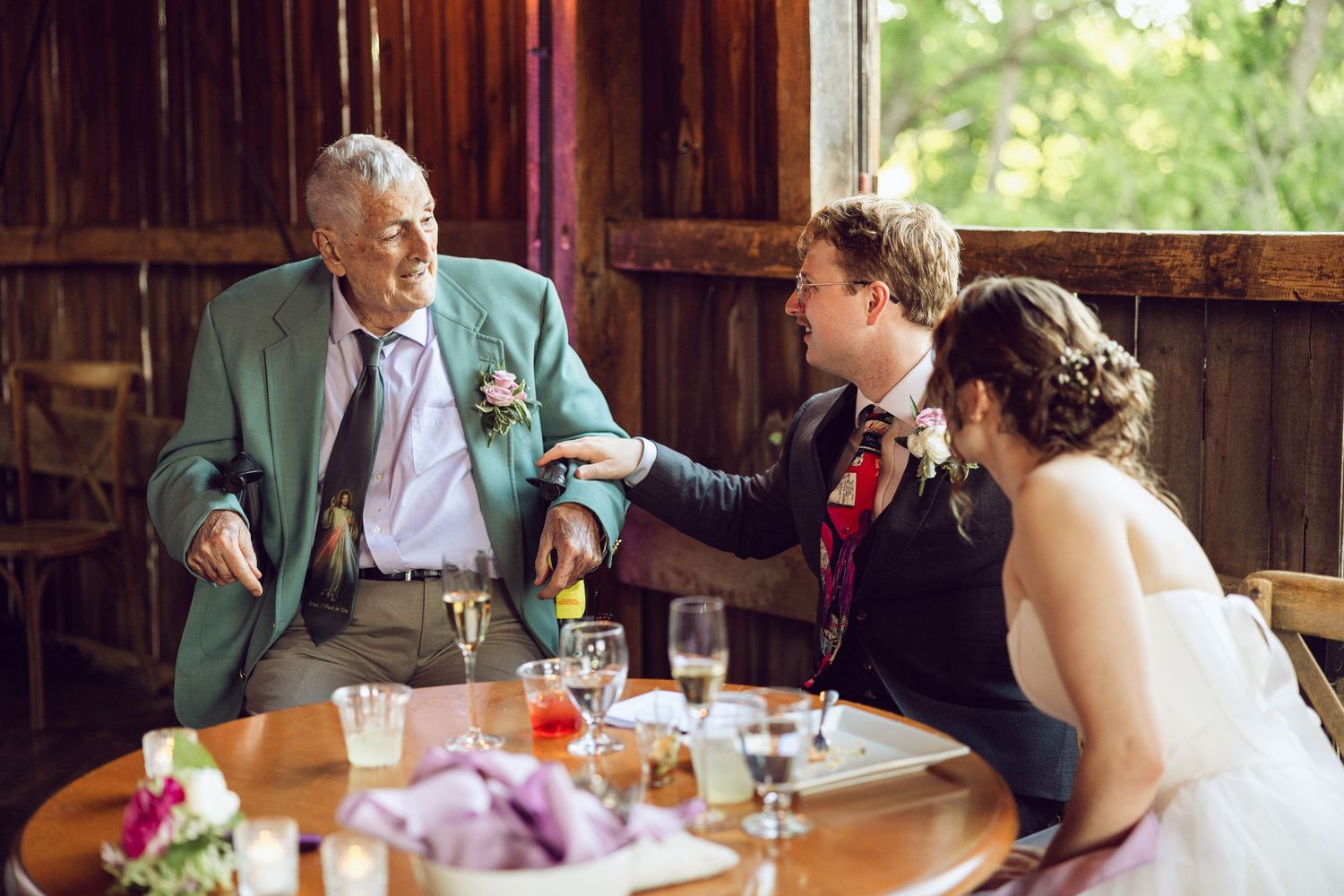 Elderly guest in a green blazer chats with the groom and bride at a wooden reception table in a rustic barn.