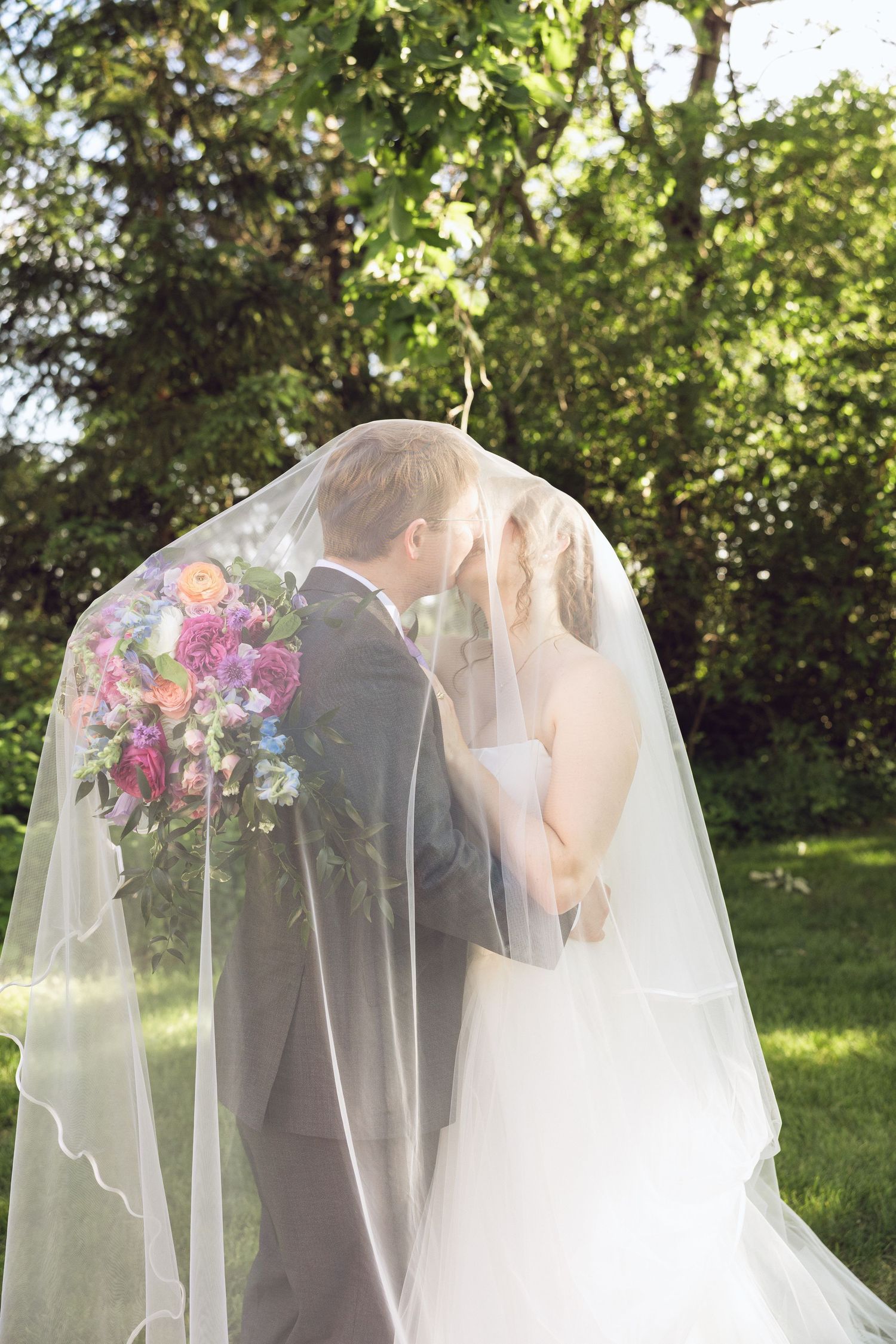 Bride and groom kiss beneath a sheer veil in a sunlit garden, with a colorful wedding bouquet resting against his back.