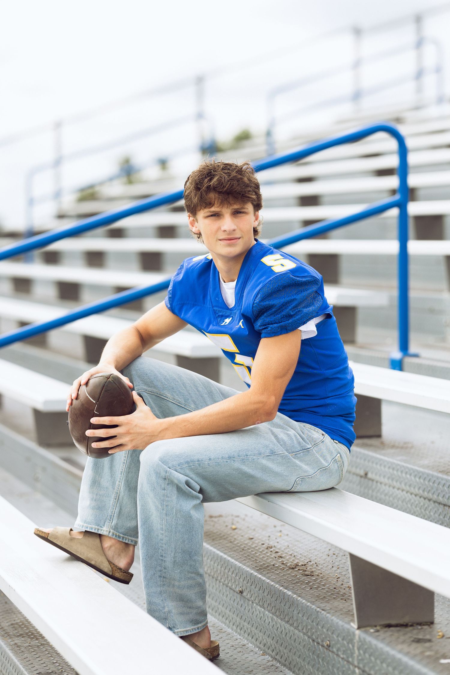 A young man in a blue football jersey holds a football while sitting on stadium bleachers with blue railings for a senior portrait.