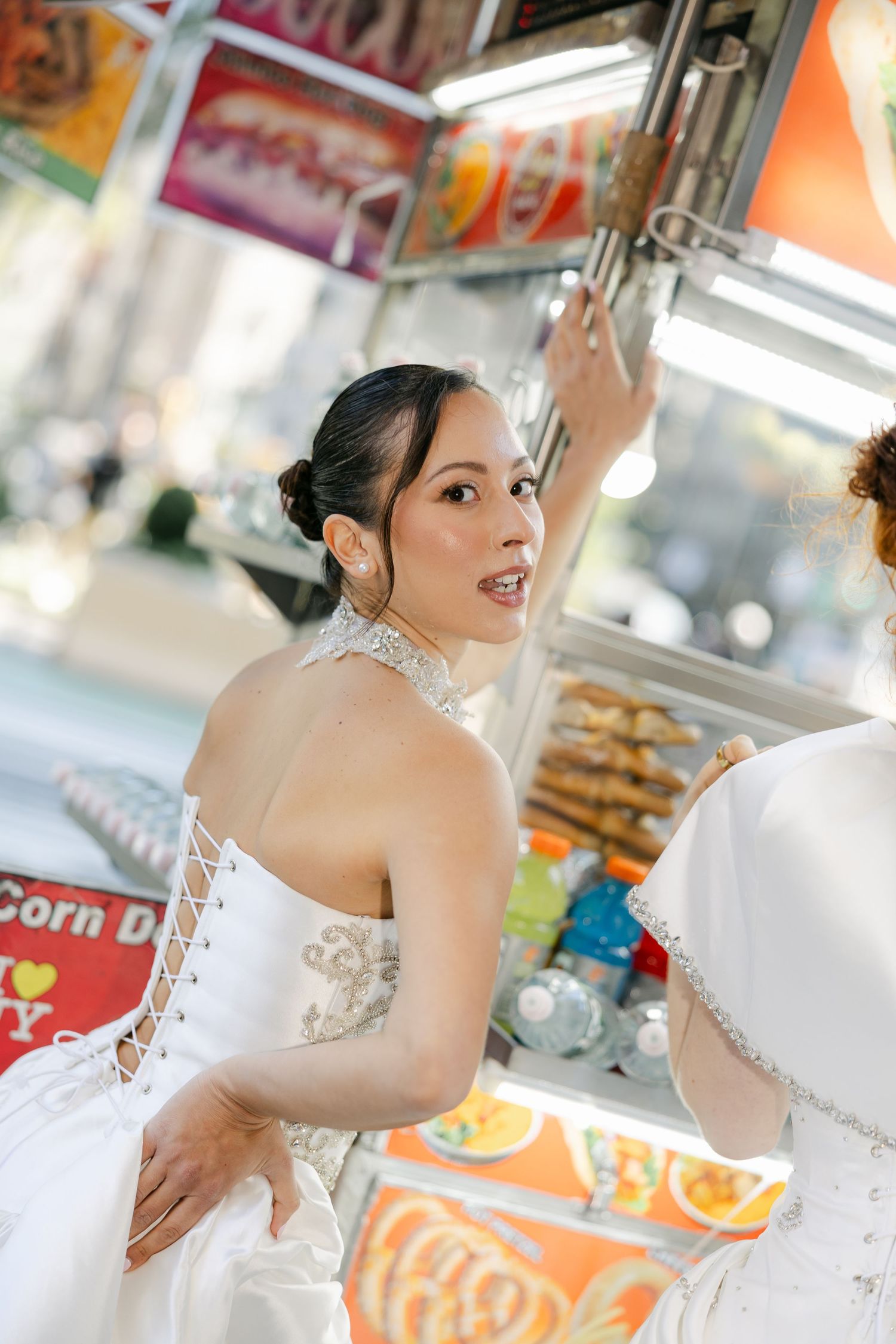 Bride in a lace-up corset-back wedding gown and crystal necklace glances over her shoulder beside a NYC food cart.