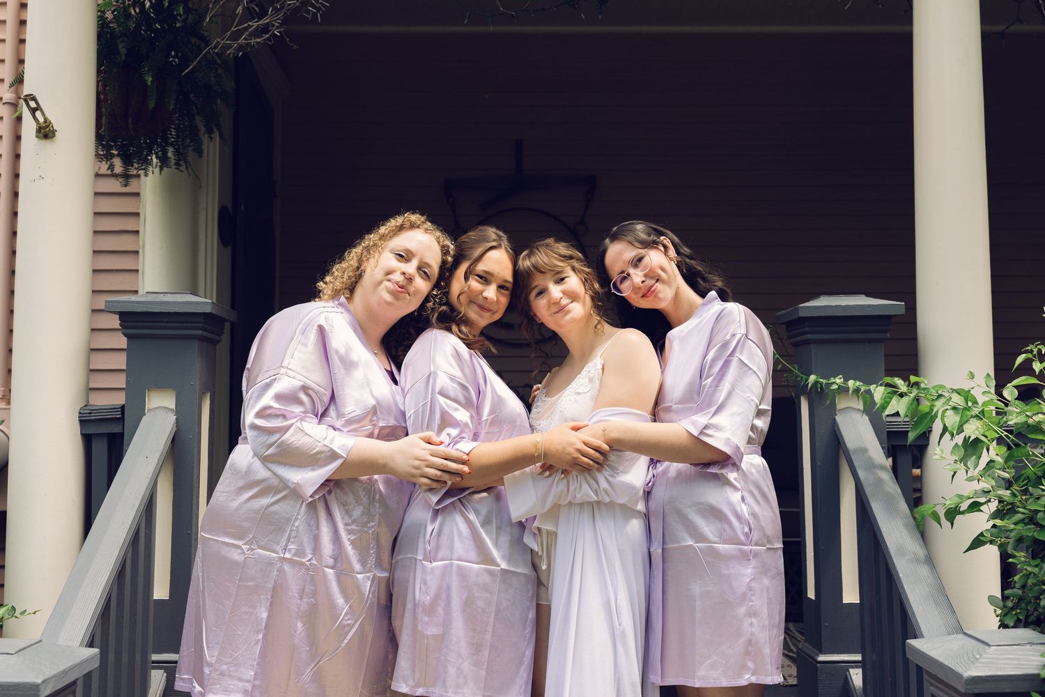 Bride and three bridesmaids in matching satin robes embrace and smile together on a porch while getting ready for the wedding.