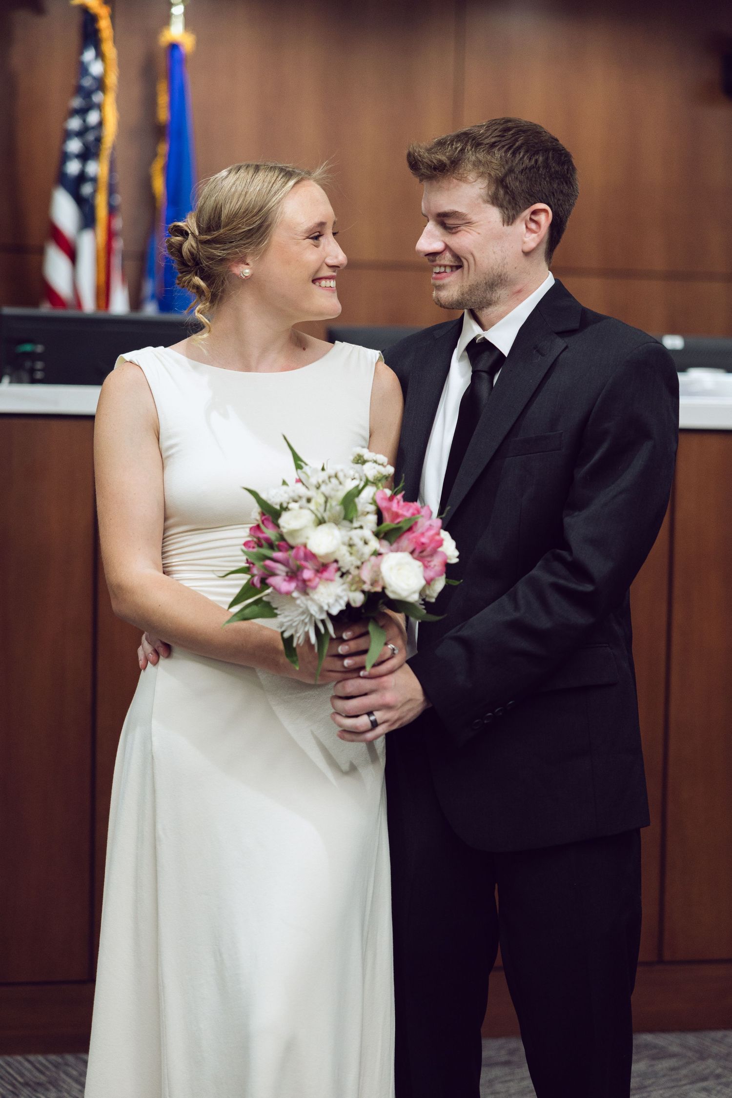 Smiling bride with a pink and white bouquet and groom in a black suit share a joyful moment in front of courthouse flags.