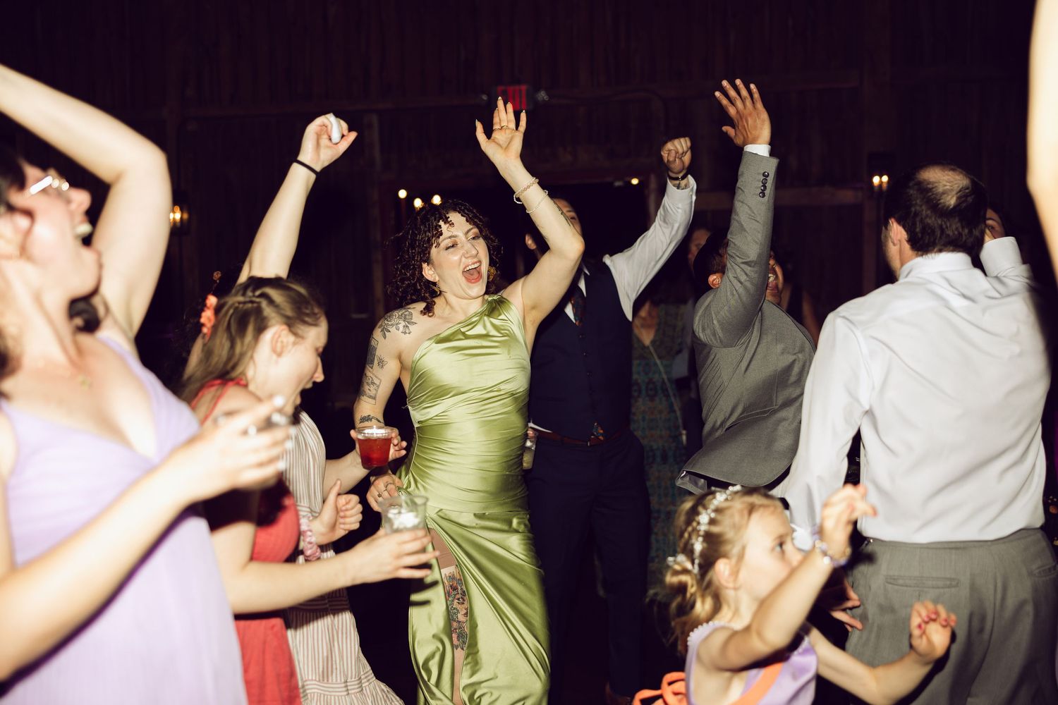 Wedding guests in colorful dresses raise their hands and dance energetically at a lively evening barn reception.