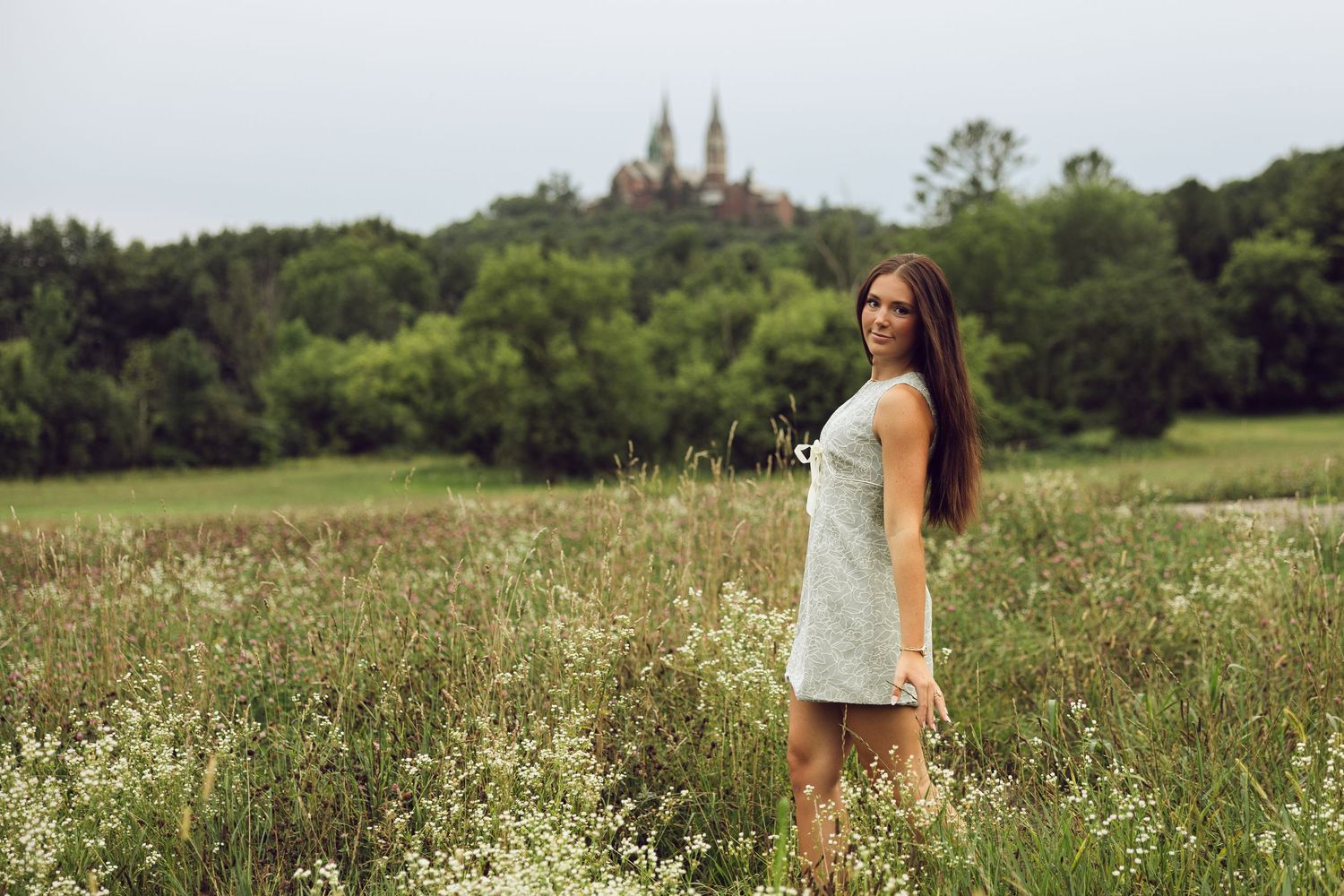 Young woman in a light dress stands in a wildflower meadow with a hilltop church and green trees in the background.