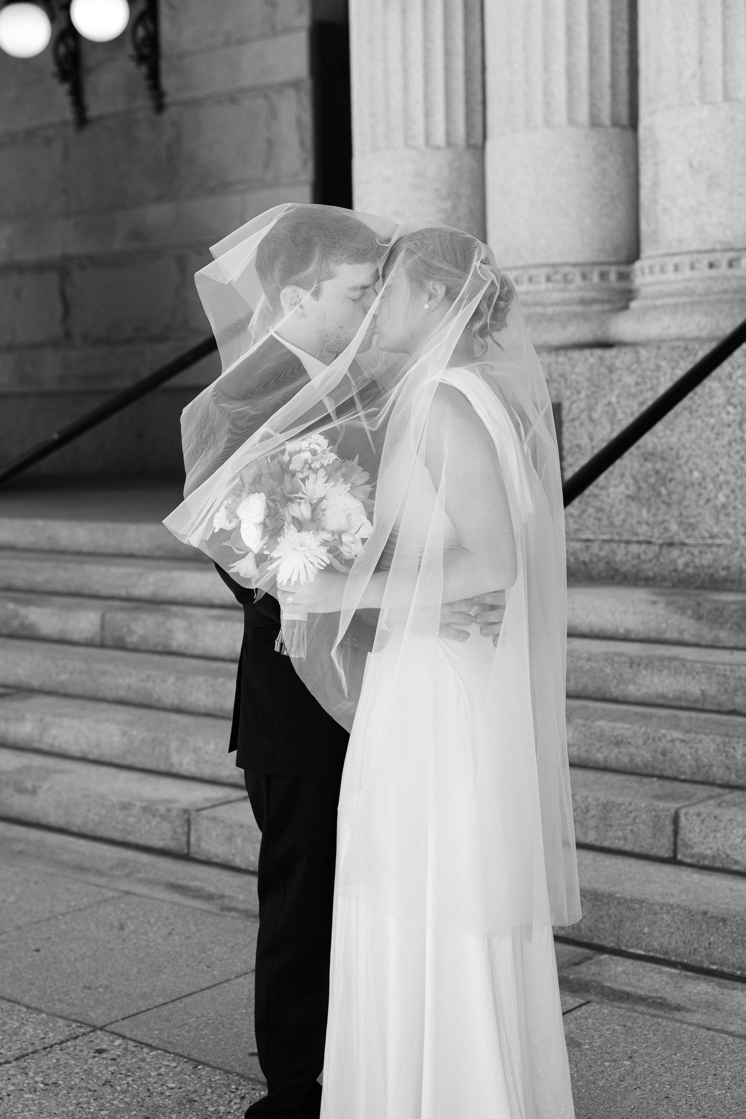 A bride and groom share a kiss beneath a sheer veil while she holds a bouquet on stone steps in a black and white photo.