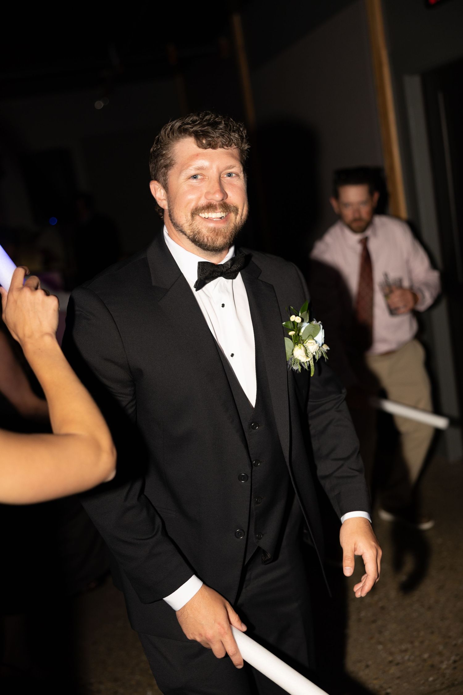 Smiling groom in a black tuxedo with bow tie and white floral boutonniere holds a glow stick on the dance floor at his reception.