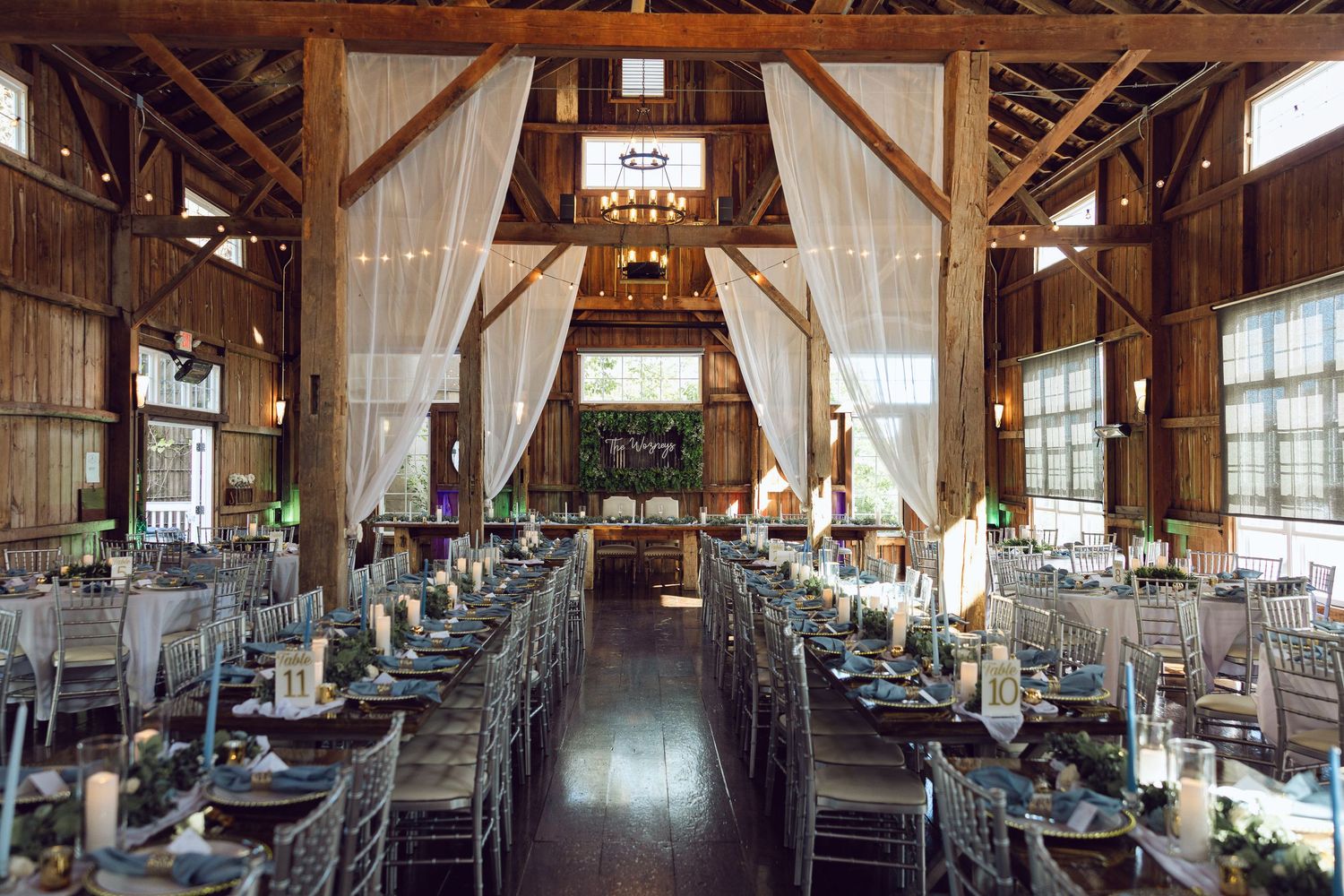 Rustic barn wedding reception hall with wooden beams, draped white fabric, and long tables set with candles and greenery.