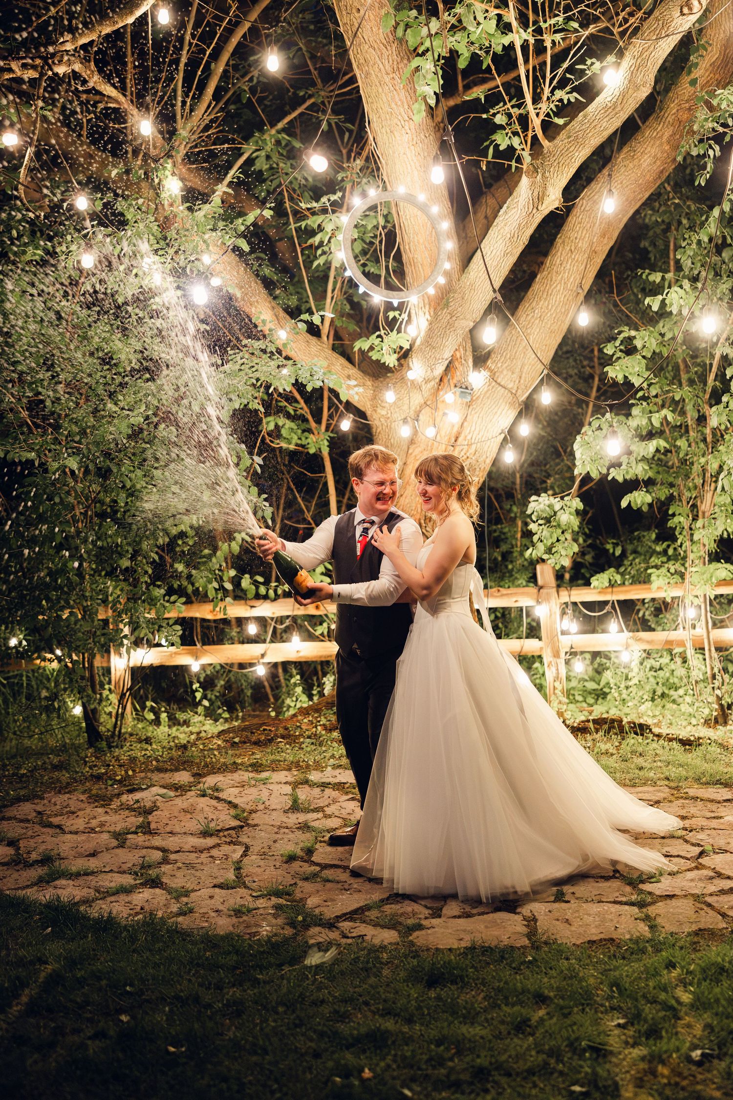 Bride and groom spray champagne beneath a large tree draped with string lights at a magical evening garden wedding.